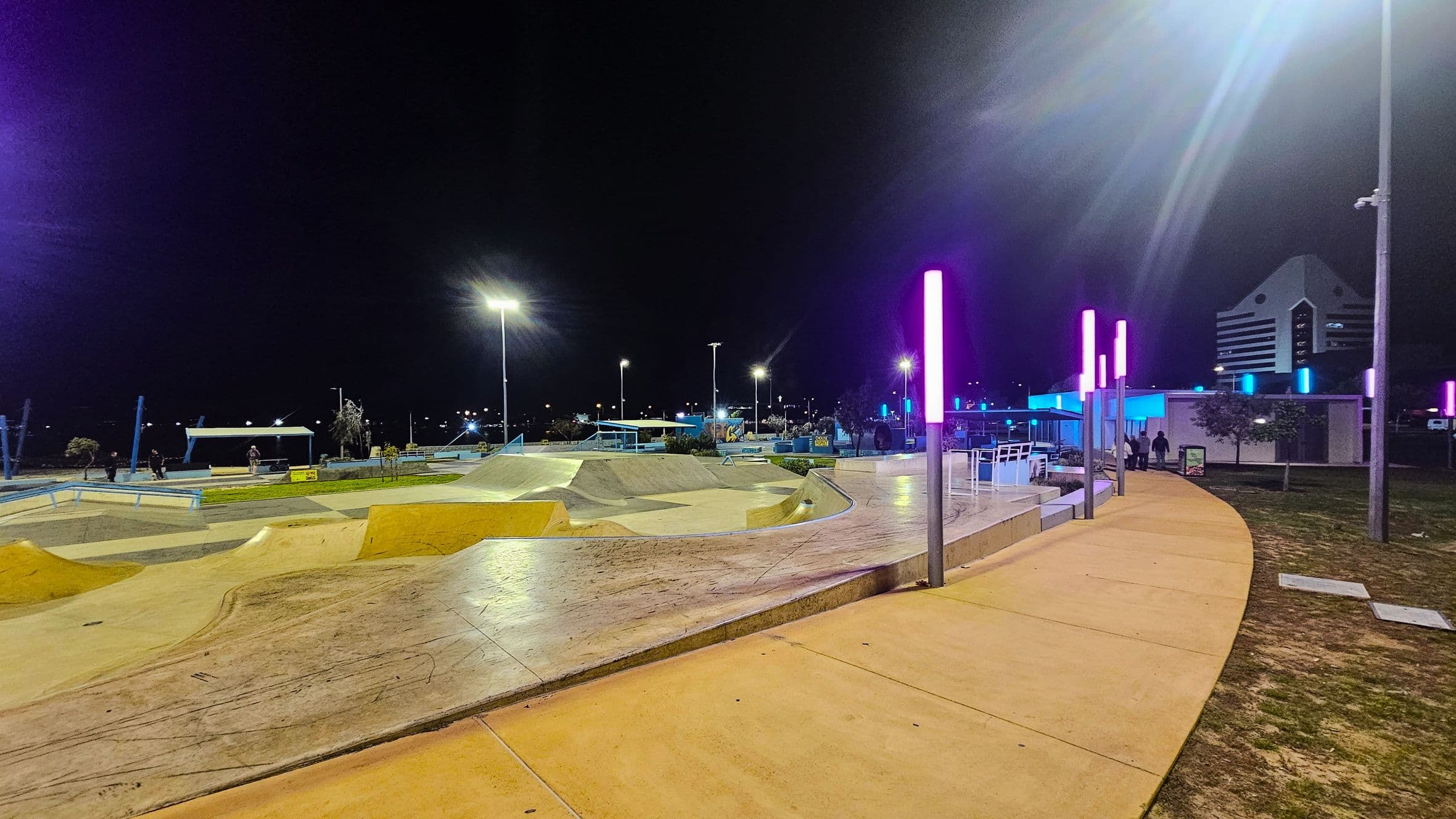 A skate park at night with various ramps and rails on the left. The ground is a mix of concrete and grass. On the right, a curved pathway is lined with tall poles featuring pink and blue lights. In the background, a modern building with a triangular roof is visible. The area is well-lit with streetlights, and a few people are present near the structures. The sky is dark, indicating nighttime.