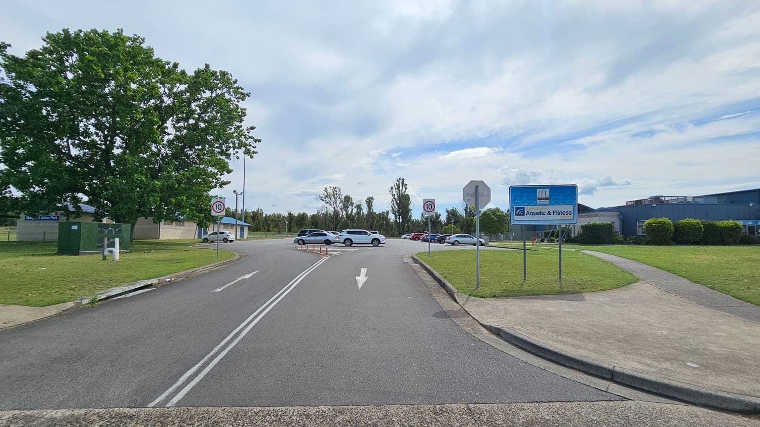 A road curves to the right with a grassy area in the centre. On the left, a car park with several vehicles is visible. A sign reads "Aquatic & Fitness" near the grassy area. In the background, a large building is partially obscured by trees and bushes. A few people are sitting on a bench near the building. The road on the right has a clear lane marking and is lined with trees and utility poles.