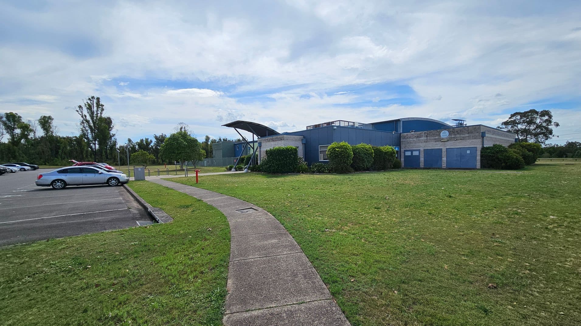 A modern building with a curved roof is on the right, surrounded by neatly trimmed bushes. A paved path leads from the foreground towards the entrance. On the left, a car park with several parked cars is visible, bordered by a grassy area. Trees line the background, providing a natural backdrop.