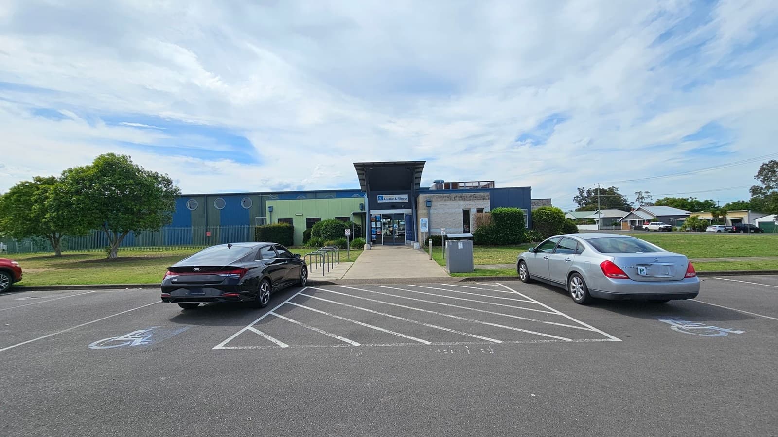 A building with a sign reading "Aquatic & Fitness" is in the centre, featuring a modern design with a mix of blue and green panels. In front, a pathway leads to the entrance, flanked by bike racks. Two cars are parked in the foreground, one black and one silver, in designated disabled parking spaces. On the left, a tree and a grassy area are visible, while on the right, there are more trees and residential houses in the background.