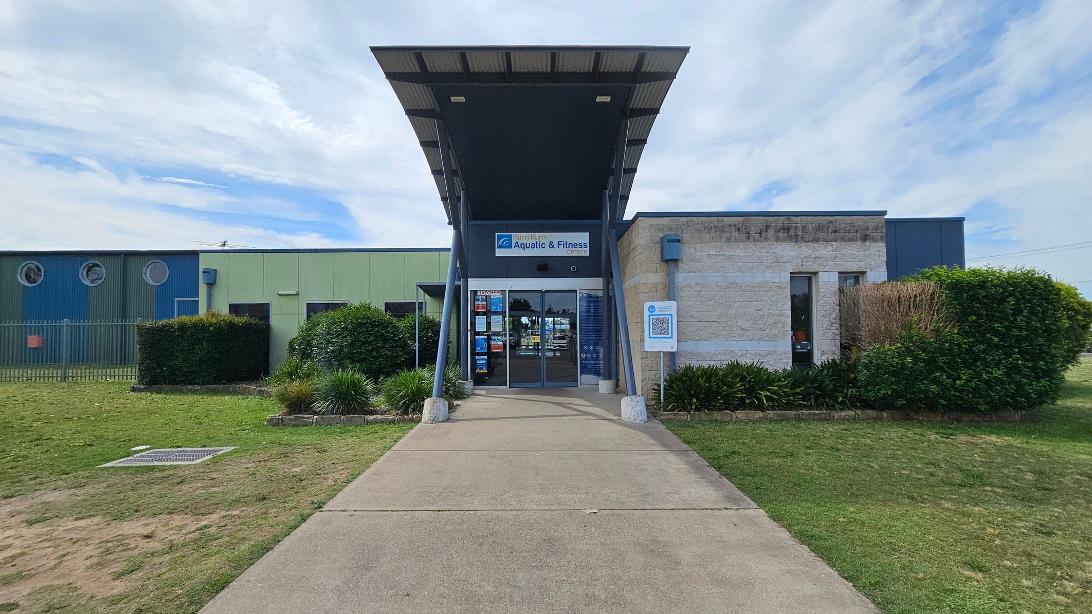 A building entrance with a modern design, featuring a large overhanging roof supported by angled metal beams. The entrance has glass doors with signs and posters. To the left, there's a blue and green wall with circular windows and a fence. On the right, a beige stone wall with narrow windows. The foreground has a concrete path flanked by grass and shrubs.
