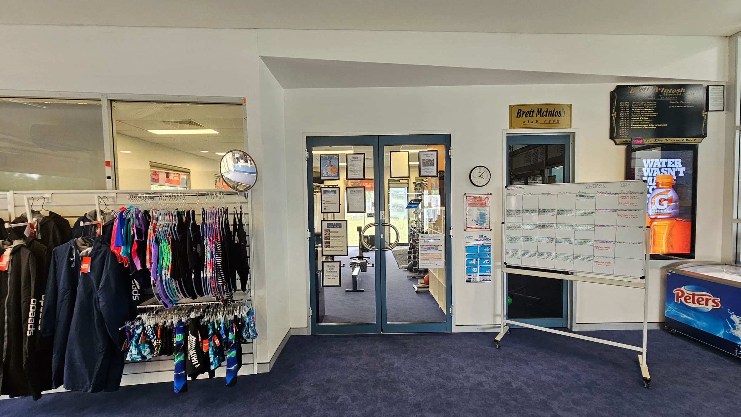 A room with a blue carpeted floor. On the left, there are racks of colourful swimwear and jackets. In the centre, double glass doors with notices lead to a gym area with equipment visible inside. A wall clock and various notices are on the wall next to the doors. On the right, a whiteboard with a calendar is displayed, and a freezer with a "Peters" logo is beneath a Gatorade advertisement. A convex mirror is mounted above the swimwear.
