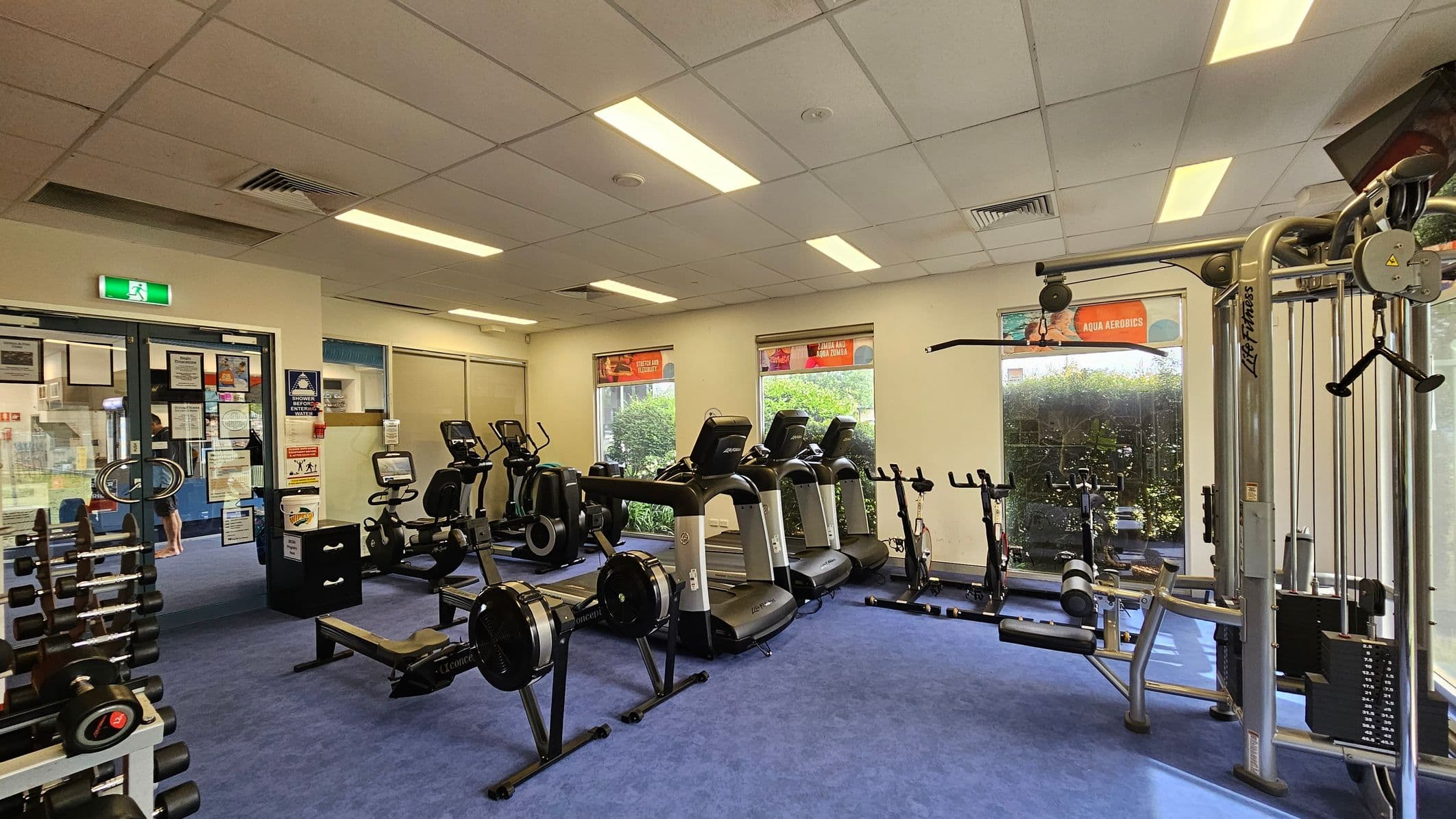 A gym with blue carpeted flooring and a white panelled ceiling. On the left, there is a rack of dumbbells and a glass door with various notices. In the centre, there are rowing machines, exercise bikes, and treadmills facing the windows. On the right, a multi-gym machine is visible. The windows have posters and offer a view of greenery outside. The room is well-lit with fluorescent lights.
