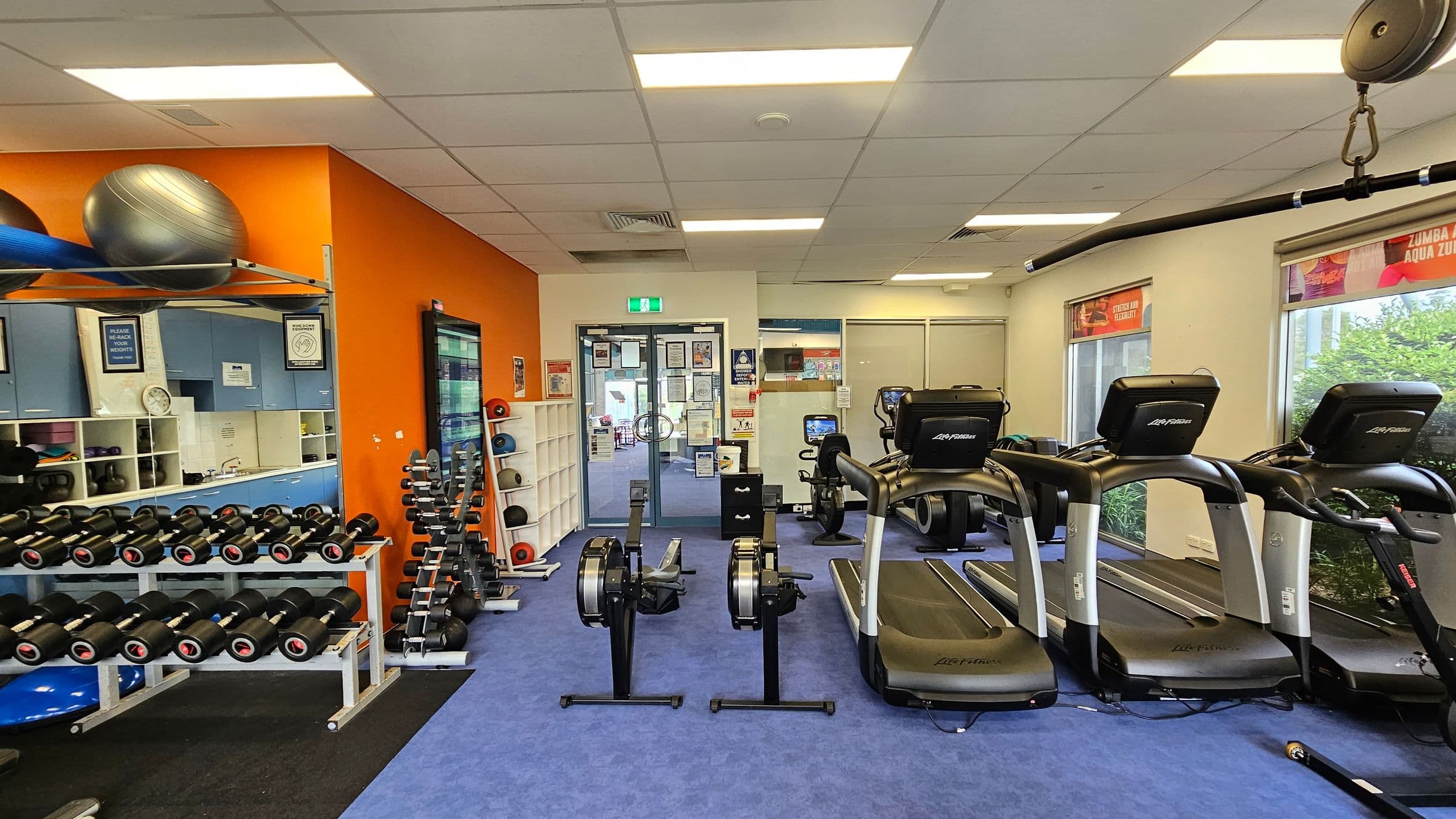 A gym with blue carpeted flooring and a white ceiling. On the left, there are racks of dumbbells and exercise balls on shelves against an orange wall. In the centre, two rowing machines face the entrance, which has glass doors and various notices. On the right, three treadmills are positioned near windows with greenery visible outside. The room is well-lit with fluorescent lights.