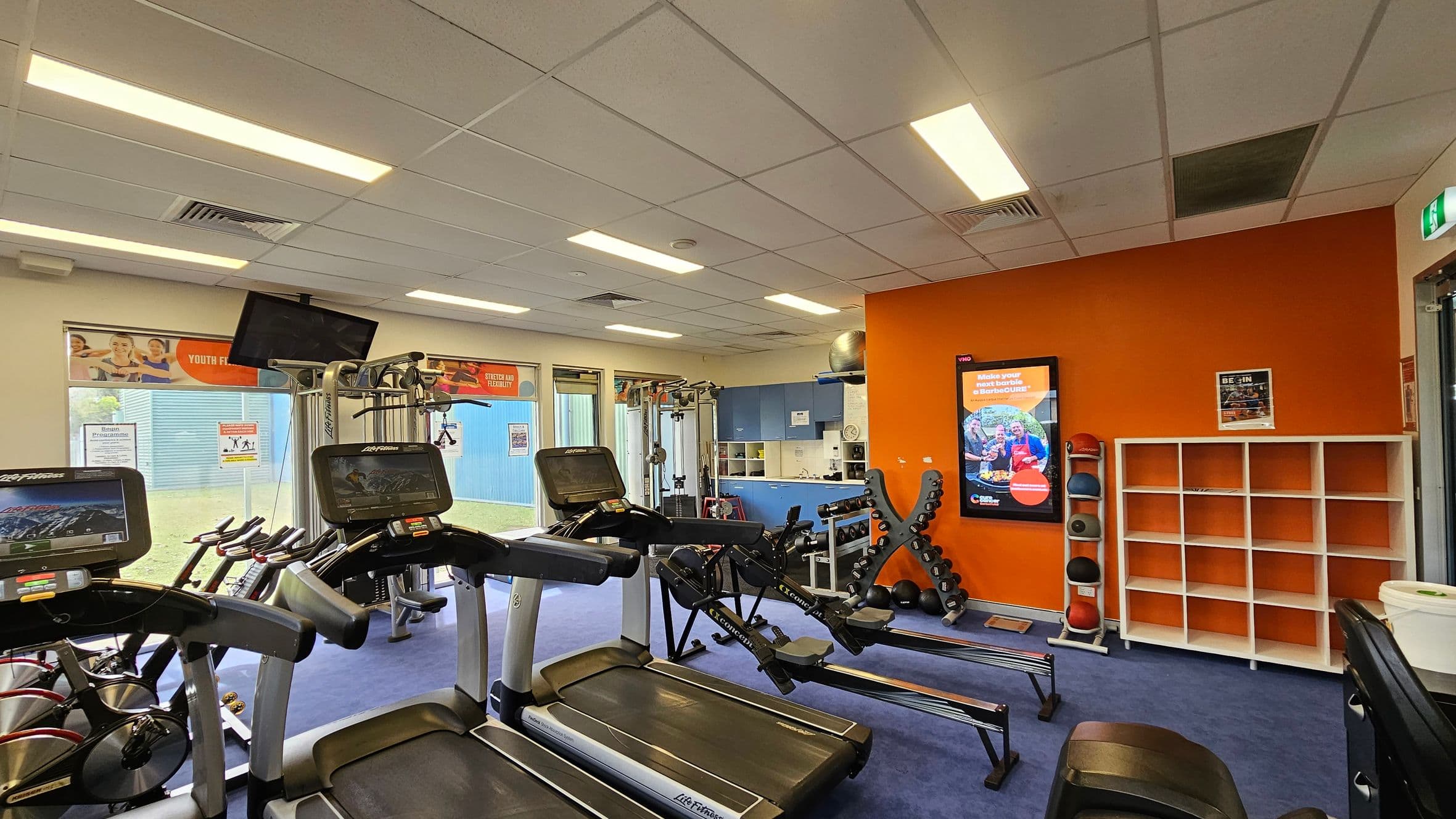 A gym room with blue carpeted flooring and a white-tiled ceiling with fluorescent lights. On the left, there are treadmills and exercise bikes with digital screens. In the middle, a rowing machine and a weight rack with dumbbells. On the right, an orange wall features a digital display and a white shelving unit. Medicine balls are stacked vertically next to the shelves. Windows with posters are in the background, allowing natural light to enter.