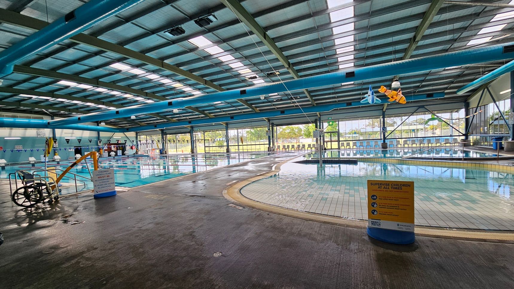 Glass doors leading to an indoor pool area. Above the doors, there is an honour roll for the Kurri Kurri Aquatic Centre. On the left, a blue sign with swimming safety tips and a QR code. On the right, a noticeboard with sections for parent and programme information. Through the doors, a pool with a yellow safety sign is visible, surrounded by a wet concrete floor. Blue chairs and a bin are also present. The ceiling has blue beams and hanging lights.
