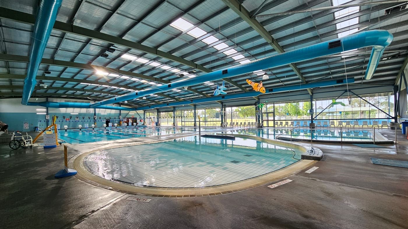 Indoor swimming pool area with a large, shallow pool in the foreground. The floor is wet and tiled around the pool. On the left, a wheelchair and a yellow pool lift are visible. The background features a larger lap pool with lane dividers and a row of blue chairs along the glass wall. The ceiling is high with exposed metal beams and blue ventilation ducts. Hanging from the ceiling are inflatable sea creatures. Large windows allow natural light to enter.