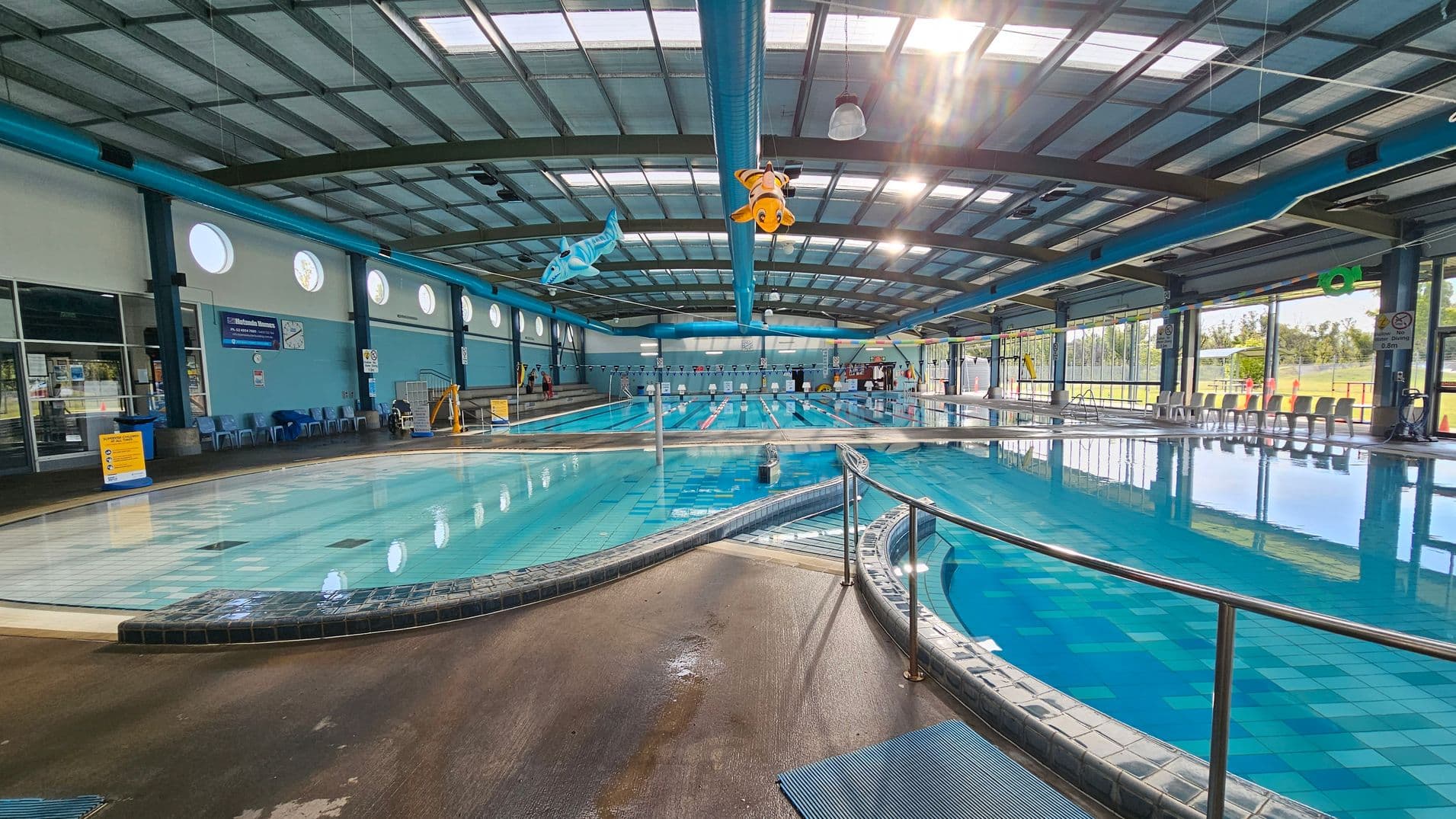 An indoor swimming pool with multiple lanes stretches across the centre. On the left, there are blue walls with circular windows and a row of chairs. Overhead, colourful fish decorations hang from the ceiling. The right side features large windows allowing natural light to illuminate the pool area. The floor is wet and tiled, with a metal railing curving around the pool's edge. The ceiling is made of metal beams with skylights.