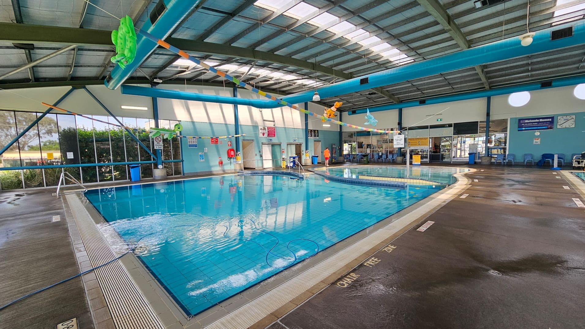 Indoor swimming pool with clear blue water, surrounded by a wet, dark brown tiled floor. On the left, large windows with greenery outside. Overhead, colourful inflatable toys hang from the ceiling. The ceiling is high with exposed beams and skylights. On the right, a row of blue chairs and a clock on the wall. Signs and safety equipment are visible along the walls. A person in an orange shirt stands near the pool's edge.