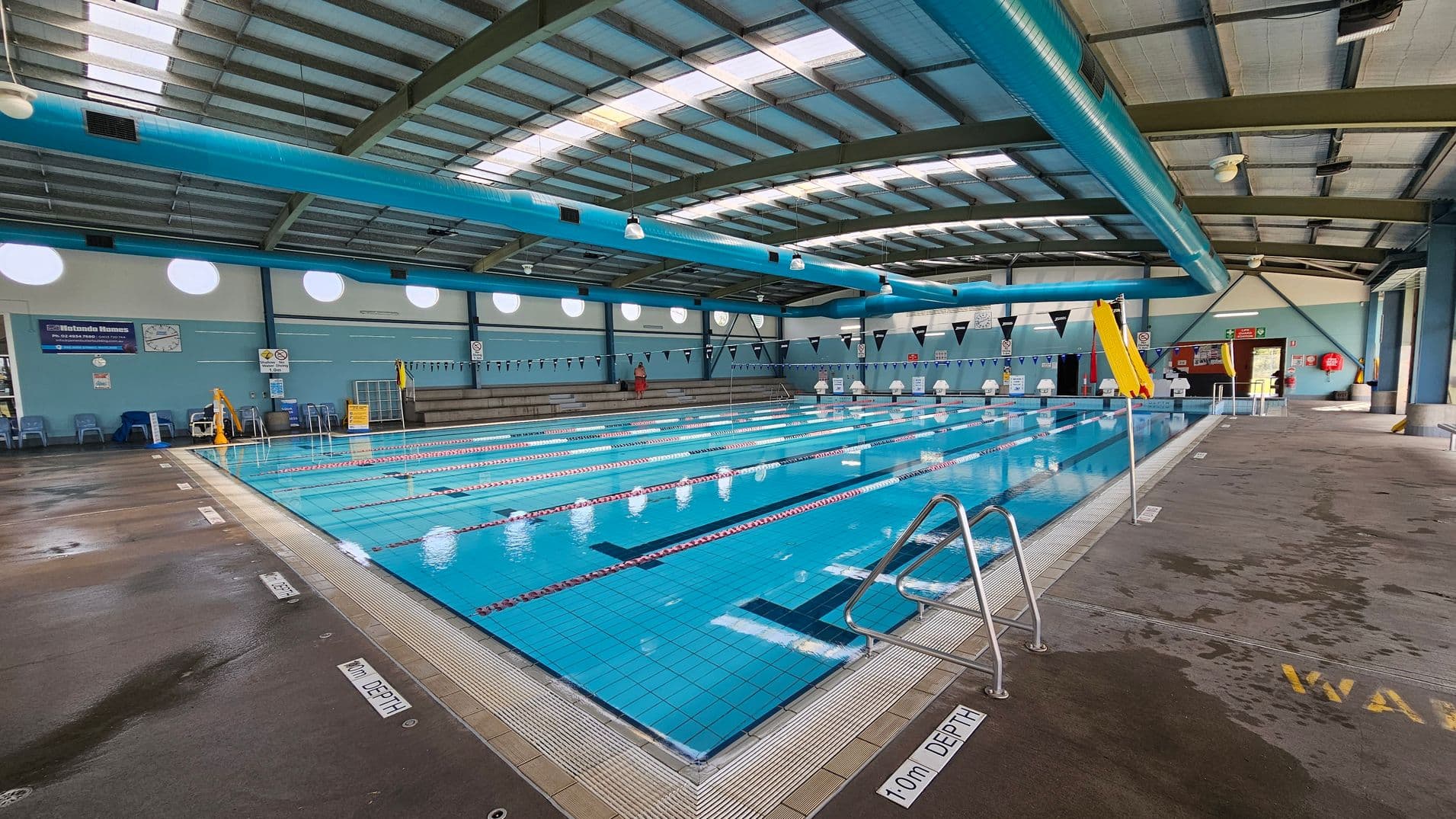 An indoor swimming pool with multiple lanes, marked by red and white lane dividers. On the right, a yellow slide is visible near the pool's edge. The pool is surrounded by a wet concrete floor with depth markers. On the left, there are blue chairs and a wall with circular windows and signs. The ceiling is high with exposed beams and large blue ventilation ducts. The far end of the pool has a lifeguard chair and more seating.