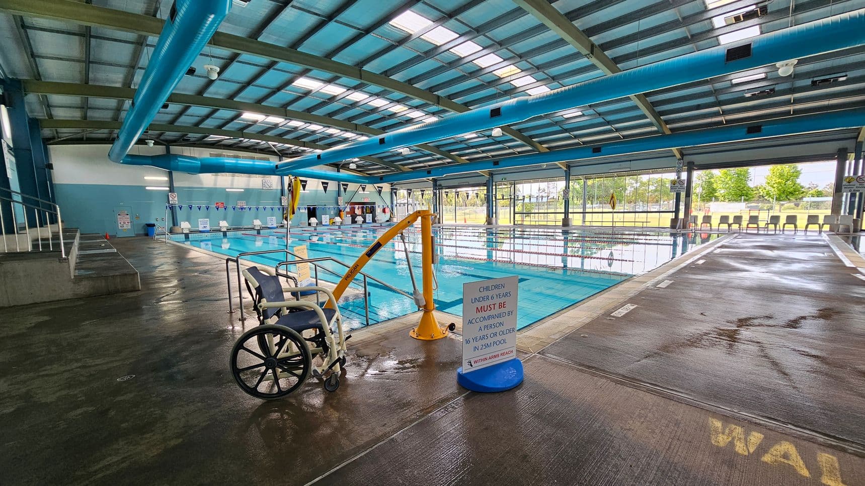 Indoor swimming pool with a wheelchair-accessible lift on the left. A sign nearby states, "Children under 6 years must be accompanied by a person 16 years or older in 25m pool." The pool is in the centre, with lanes marked by floating dividers. On the right, there are several chairs lined up against large windows that let in natural light. The ceiling is high with blue ventilation ducts, and the floor is wet, indicating recent use.