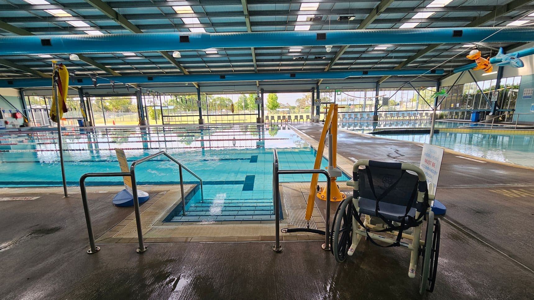 Indoor swimming pool with lanes marked by ropes. On the right, a wheelchair is positioned near a pool lift. The floor is wet and made of non-slip material. Large windows in the background provide a view of trees and grass outside. The ceiling is high with exposed beams and lighting. A yellow flag is on the left, and colourful pool toys hang from the ceiling.