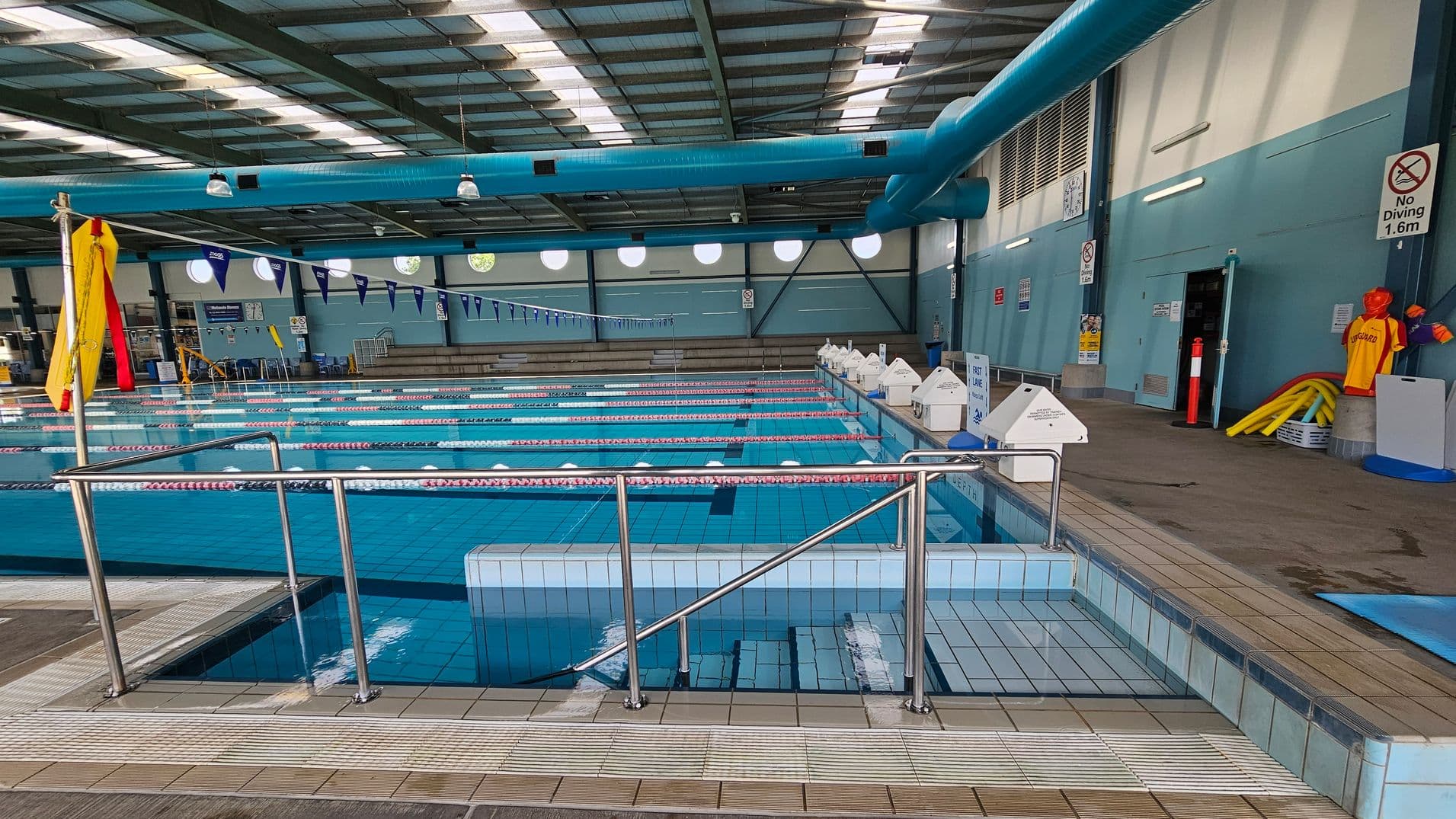 Indoor swimming pool with multiple lanes, separated by red and white lane dividers. On the right, a wall with "No Diving" signs and lifeguard equipment, including a mannequin wearing a lifeguard shirt. The left side has a metal railing and a small step leading into the pool. The ceiling features large blue ventilation ducts and overhead lights. The floor is tiled, and there are circular windows along the back wall.