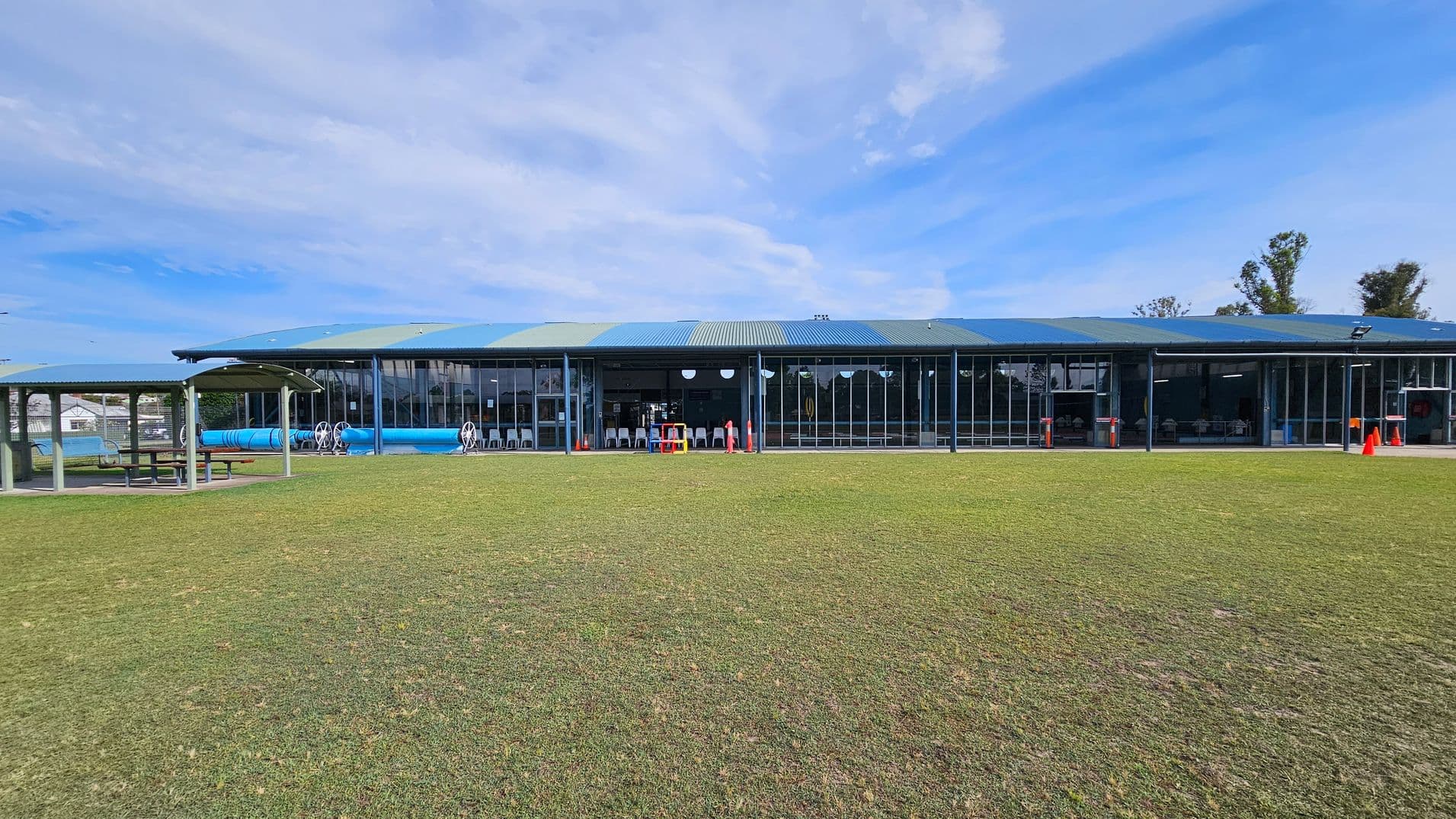 A large, single-storey building with a blue and green roof and glass walls stretches across the background. In front of the building, there are several white chairs and a few orange cones. To the left, a covered picnic area with a table and benches is visible. The foreground is a grassy field with patches of brown. A few trees are visible in the distance on the right.