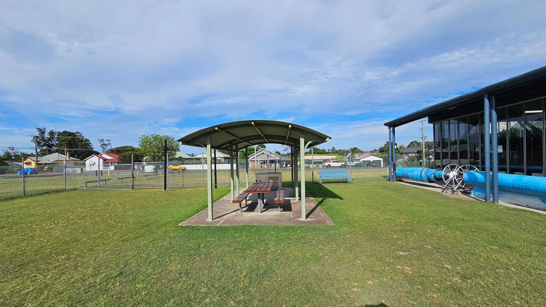 A grassy area with a covered picnic table and benches in the centre. To the right, there's a building with large windows and a blue cylindrical structure, possibly a pool cover roller. In the background, there are several houses and a fence enclosing the area.