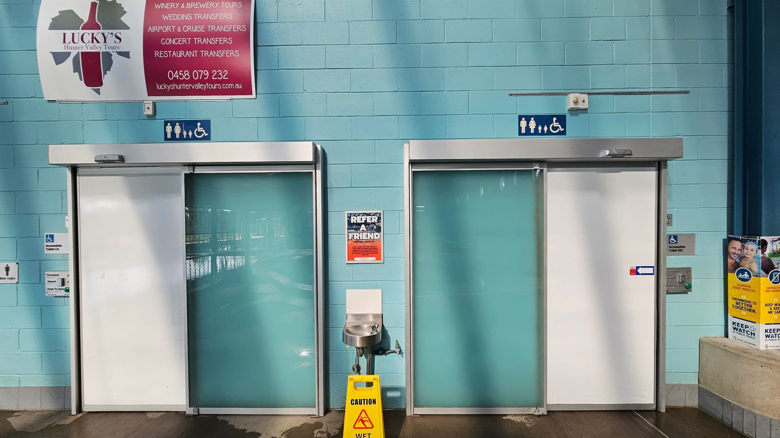 Two accessible toilet doors with sliding panels are set in a light blue brick wall. Above the doors are signs indicating accessibility. Between the doors is a drinking fountain with a yellow "Caution Wet" sign on the floor. A red and white advertisement is mounted on the wall above. To the right, a poster with safety information is visible. The floor is tiled, and the ceiling is not shown.