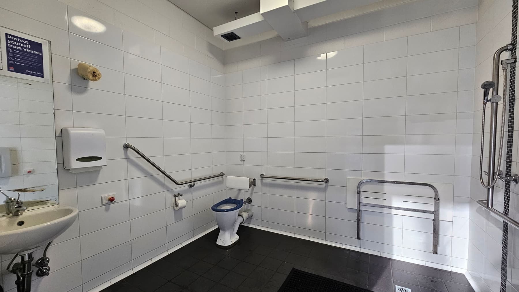 A public accessible bathroom with white tiled walls and a black tiled floor. On the left, there is a small sink with a soap dispenser and a paper towel holder above it. A sign about virus protection is visible. In the centre, a toilet with a blue seat is equipped with support rails on both sides. On the right, there is a foldable shower seat and a handheld showerhead attached to the wall. The ceiling has a ventilation duct.