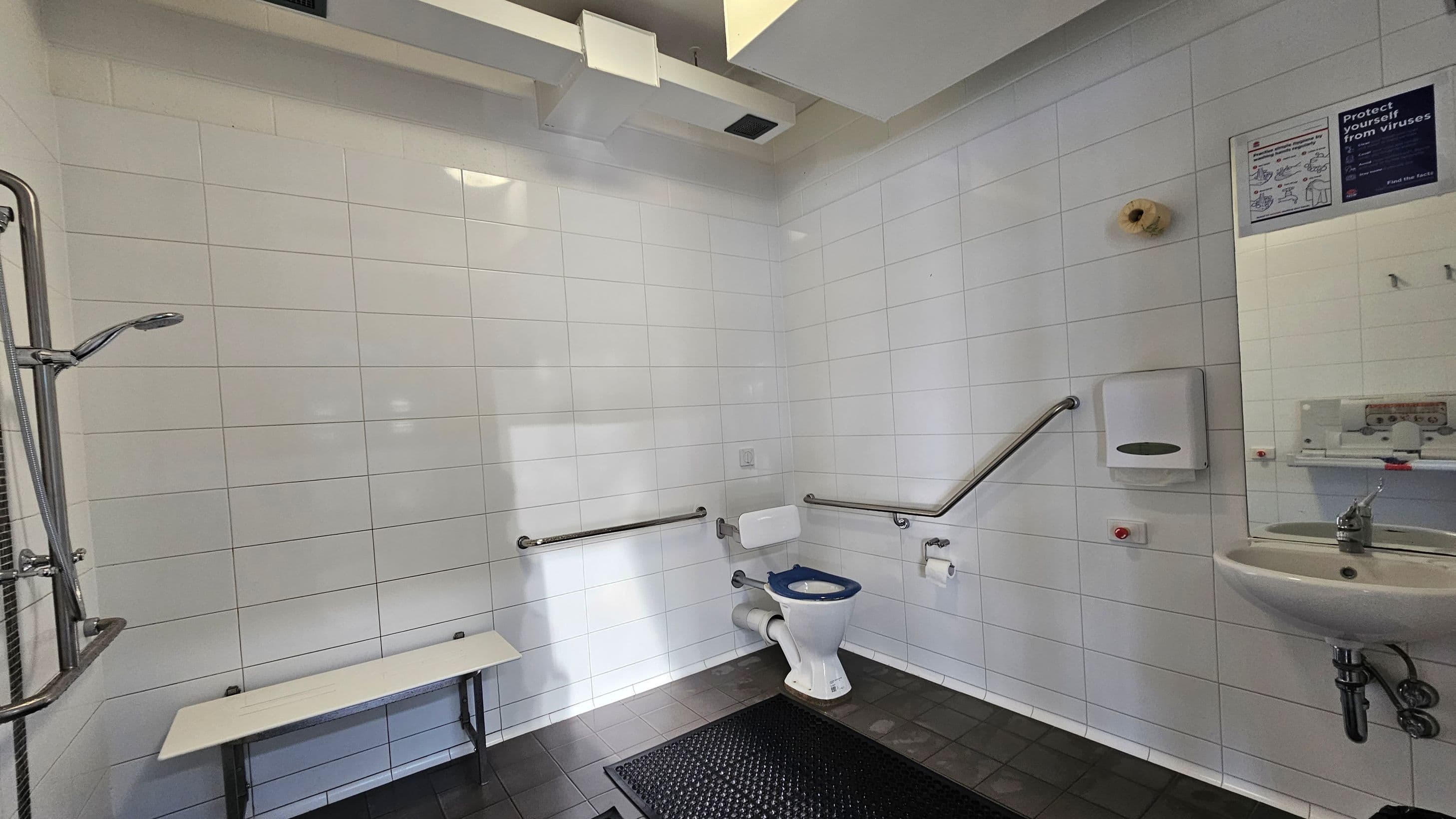 A bathroom with white tiled walls and a dark tiled floor. On the left, there's a shower with a metal showerhead and a foldable bench. In the centre, a toilet with support rails on both sides. On the right, a sink with a mirror above it, a soap dispenser, and a paper towel holder. A sign with hygiene instructions is visible on the wall. The ceiling has a vent and a light fixture.