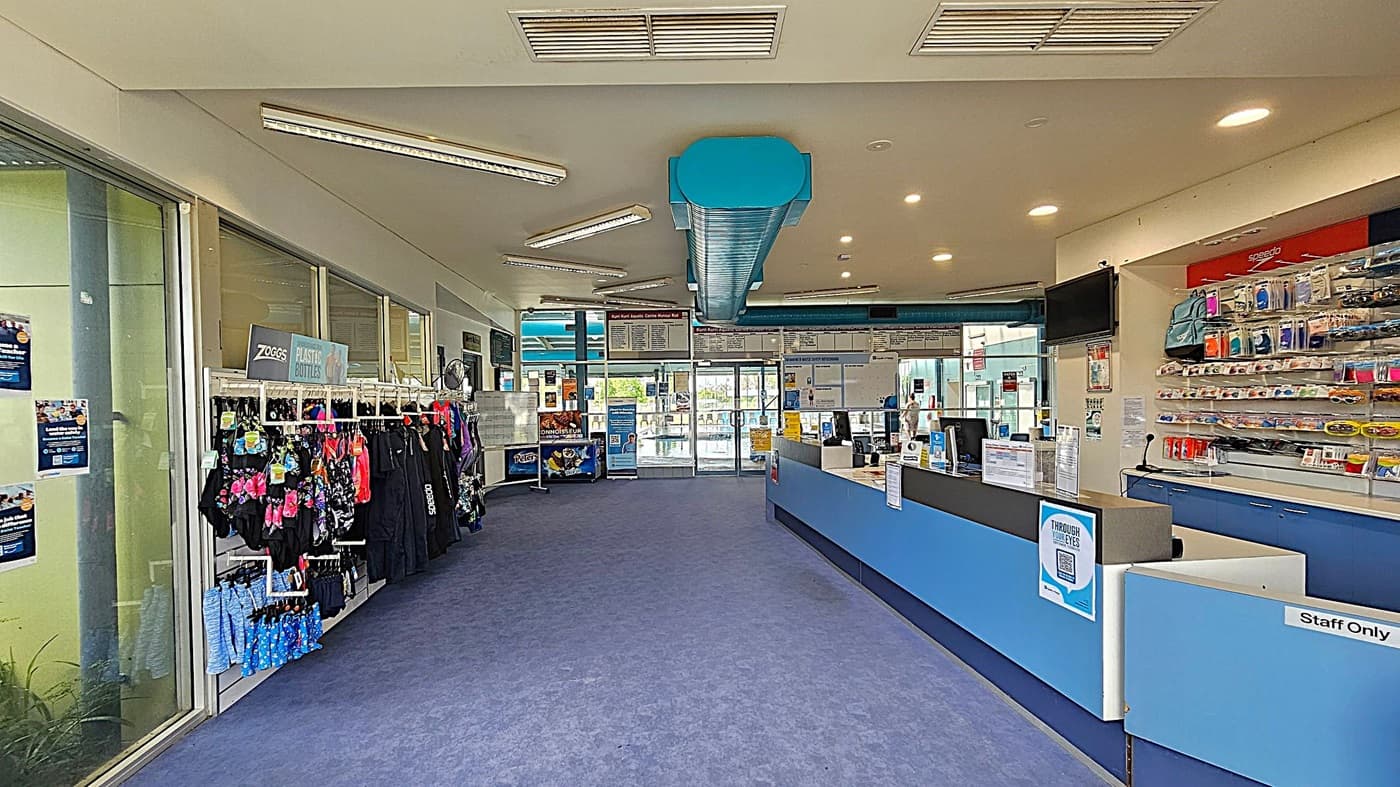 Glass entrance with sliding doors. On the left, several posters and notices are displayed, including a "HAZCHEM" sign. On the right, a large blue sign lists "CONDITIONS OF ENTRY" with various rules and symbols. The interior is dimly lit, showing a glimpse of a pool area in the background. The exterior wall on the right is made of beige brick, and there are blue metal columns on both sides. The floor is concrete outside and carpeted inside.