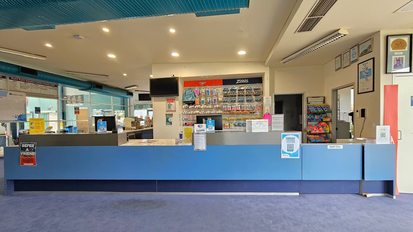 A reception area with a long blue counter in the foreground, featuring signs and a "Staff Only" label. Behind the counter, shelves display various swimming accessories like goggles and swim caps. To the right, a rack holds snacks or small items. The background includes large windows, a wall with framed certificates, and a door. The ceiling has recessed lighting and visible air ducts, while the floor is carpeted in blue.