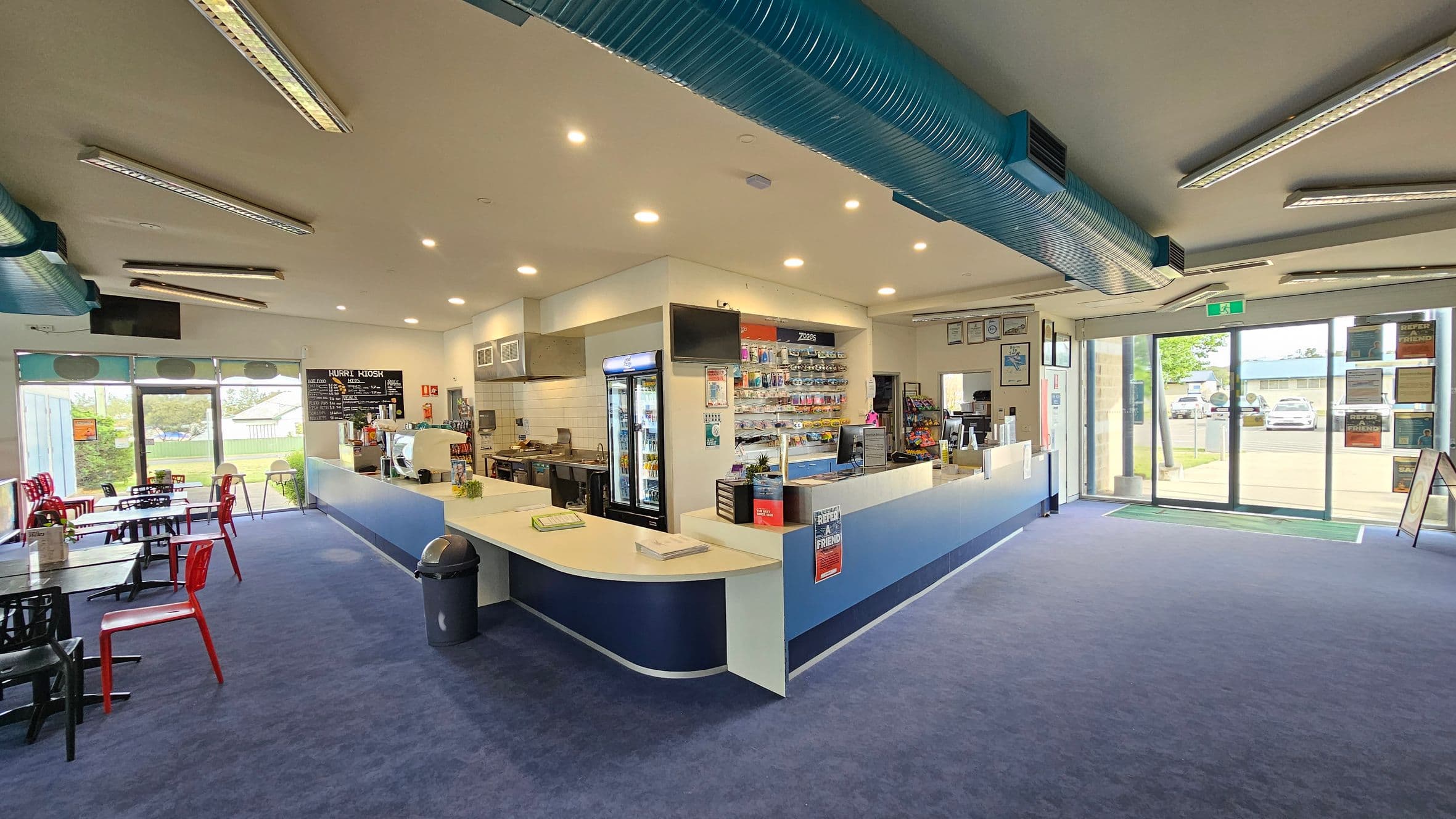 A spacious indoor café with a blue carpeted floor. On the left, there are tables and chairs, some red and some black, near large windows showing a grassy outdoor area. In the centre, a counter with a display of snacks and a fridge with drinks. The ceiling has multiple light fixtures and a prominent blue ventilation duct. On the right, glass doors lead outside, with a view of parked cars and a building. The walls have various signs and a menu board.