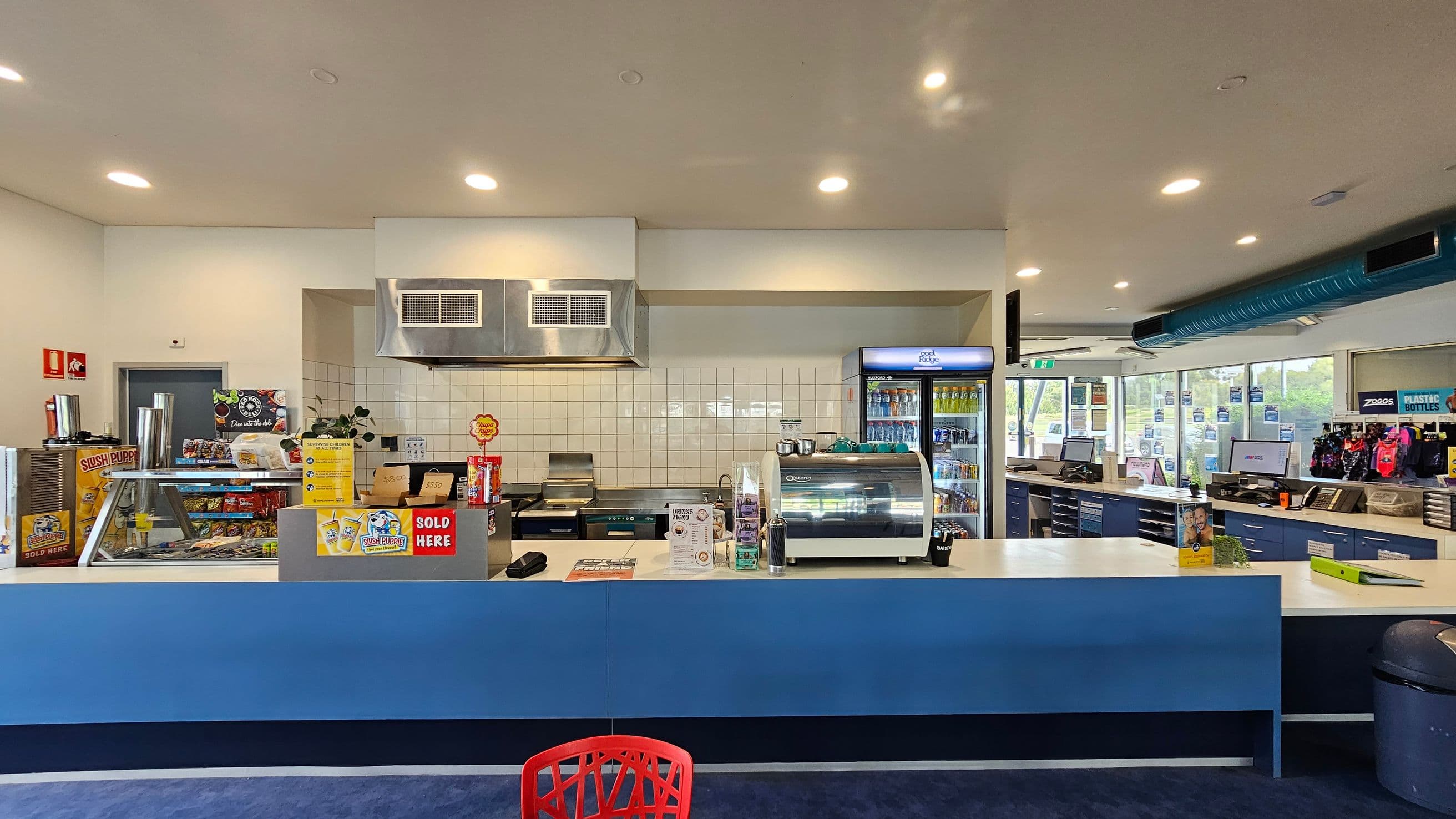 A café counter with a blue front. On the left, there is a Slush Puppie machine and a display of snacks. In the centre, a coffee machine and a menu are visible. Behind the counter, a tiled wall with a stainless steel ventilation hood and a grill. To the right, a fridge stocked with drinks and a cash register area with merchandise. The ceiling has recessed lighting, and the floor is carpeted. A red chair is partially visible in the foreground.