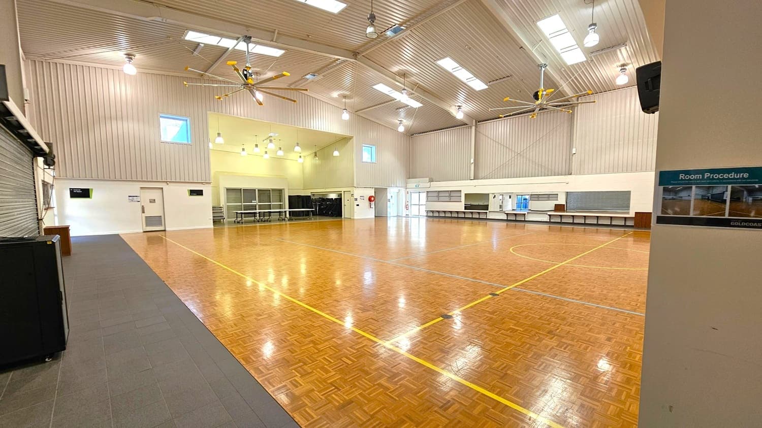 A large indoor hall with a polished wooden floor marked with yellow lines. The ceiling is high with skylights and large fans. On the left, there is a closed roller door and a small door with a sign above it. The right side has a long counter with a shutter and a sign that reads "Room Procedure." The back wall features windows and a fire extinguisher. The lighting consists of multiple hanging lamps.