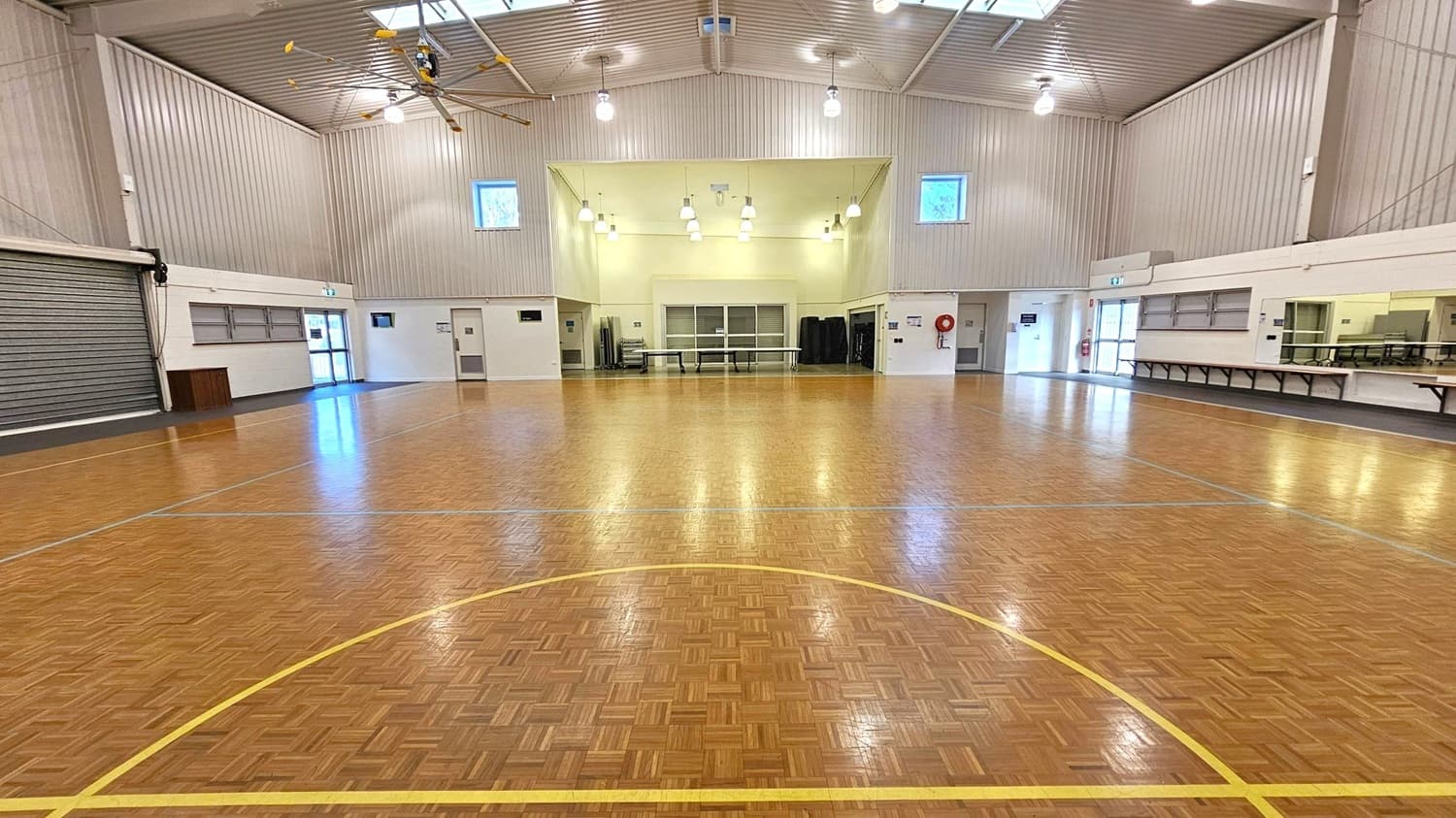 A spacious indoor hall with a polished wooden floor marked with yellow lines. The ceiling is high with exposed beams and several hanging lights. On the left, there is a large roller door and a small entrance. The back wall features two small windows and a central area with tables and stacked chairs. On the right, there are long tables and a serving area with a counter and windows. The walls are light-coloured, and the space is well-lit.