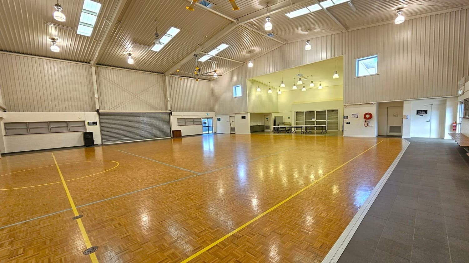 A spacious indoor sports hall with a polished wooden floor marked with yellow and blue lines. The ceiling is high with exposed beams and several skylights, along with hanging lights. On the left, there is a large roller door and a small exit door. The right side features a fire hose reel and a couple of doors, one with a sign. The back wall has a few windows and a section with tables and chairs. The floor transitions to a grey tiled area on the right.