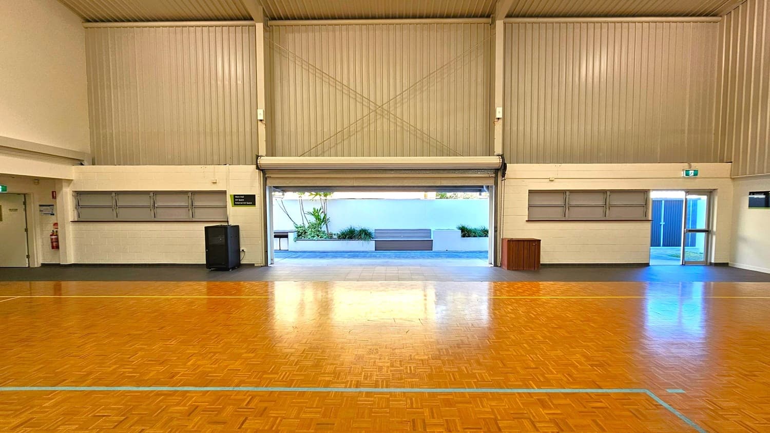 A large indoor space with a polished wooden floor and high corrugated metal walls. In the centre, a wide roller door is partially open, revealing an outdoor area with a white wall, plants, and a bench. On the left, there is a closed door and a fire extinguisher. On the right, a glass door leads outside. The ceiling is high and ribbed, and there are two sets of horizontal windows on either side of the roller door.