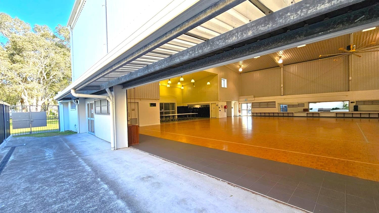 A large indoor space with a wooden floor and high ceiling, featuring multiple hanging lights and a large ceiling fan. The room is open to the outside through a wide, raised garage-style door. Outside, there is a concrete path leading to a metal gate, with trees visible in the background. The interior walls are light-coloured, and there are several windows and a door on the far wall. Tables are lined up against the wall inside.