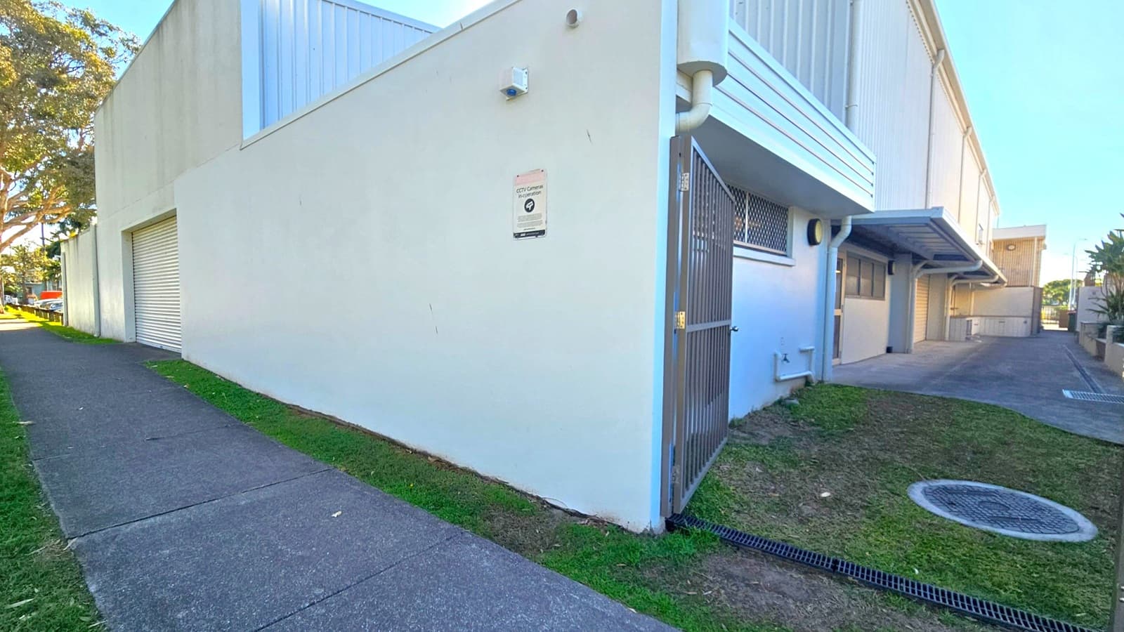 A large, light-coloured building with a flat roof and a roller door on the left side. A CCTV sign is mounted on the wall near a metal gate. The building has a small window with a security grille. A concrete path runs alongside the building, bordered by grass and a few trees. On the right, there is a narrow driveway leading to the back of the building, with a covered area and additional doors. The ground is a mix of concrete and grass.