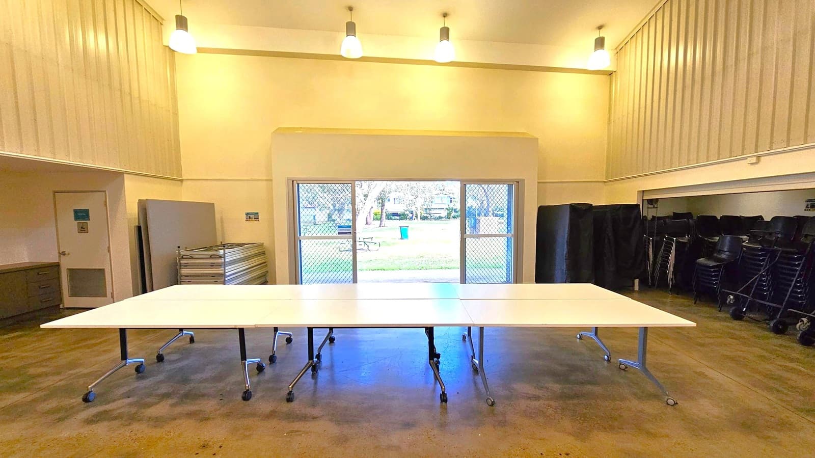 A room with a high ceiling and three pendant lights. In the centre, there are several white tables on wheels arranged together. The floor is a polished concrete surface. On the left, there is a stack of folded tables and a door. On the right, there are stacked chairs. The back wall features a large window with a mesh screen, offering a view of a grassy area with trees, a picnic table, and a blue bin.