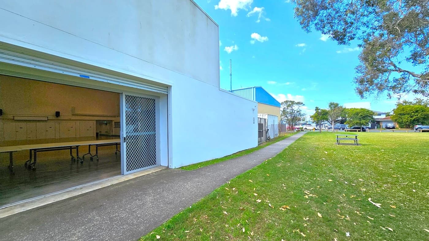 A building with a partially open roller door and a mesh screen is on the left, revealing tables inside. A concrete path runs alongside the building, leading towards the background where more buildings and parked cars are visible. On the right, a grassy area with scattered leaves and a picnic table is bordered by trees. The sky is clear and blue.
