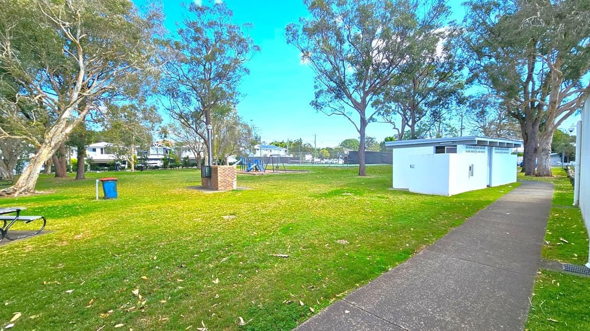 A park with a grassy area and scattered trees. On the left, there's a blue bin and a picnic table. In the centre, a playground with swings and a brick barbecue area. On the right, a white public restroom building is adjacent to a paved path. In the background, residential houses are visible beyond a fence.