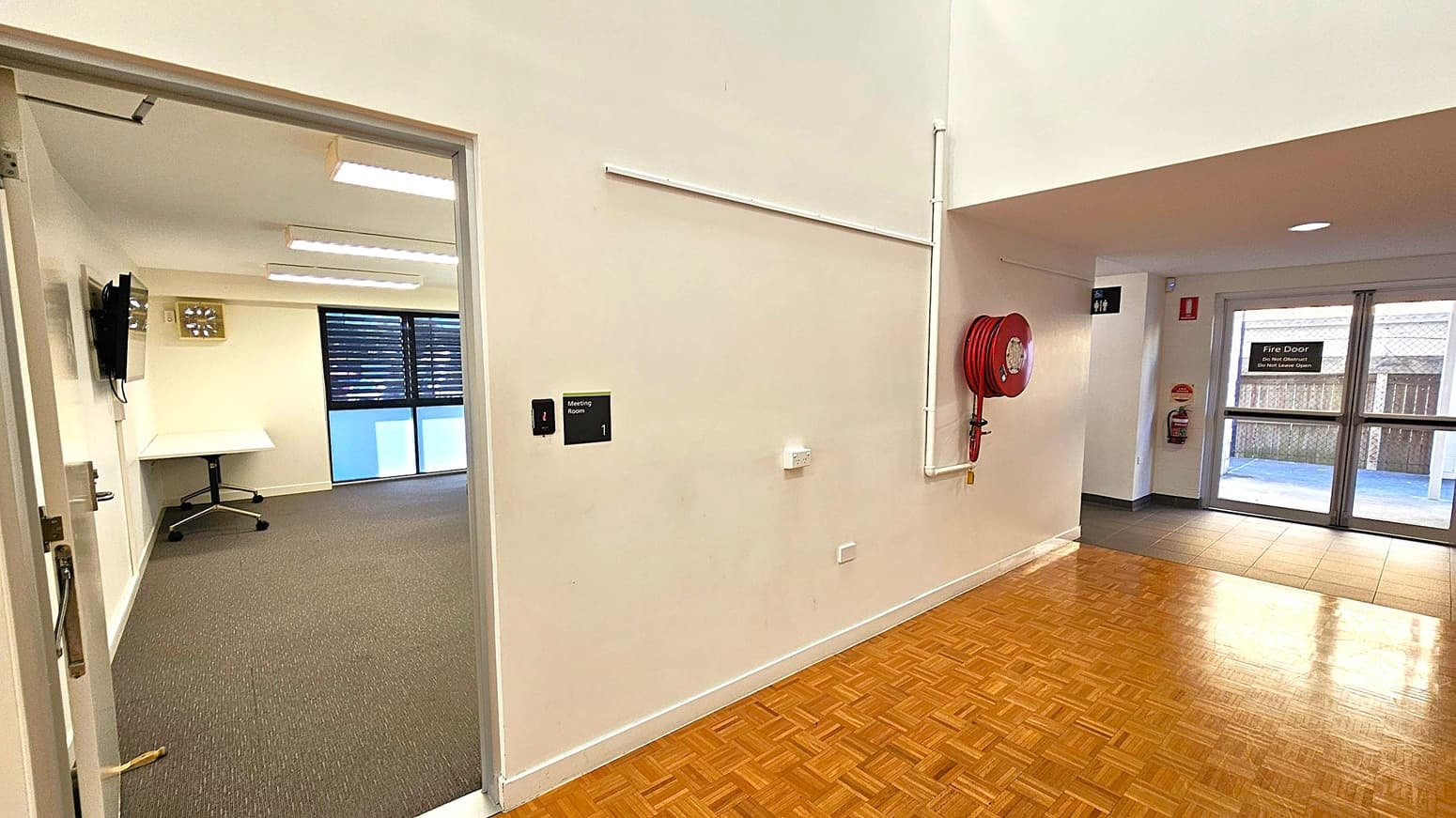 A hallway with a parquet floor and a white wall. On the left, an open door leads to a meeting room with a carpeted floor, a wall-mounted TV, and a desk. The right side features a red fire hose reel mounted on the wall. In the background, there is a fire door with a sign, and a tiled floor area leading to the outside. Ceiling lights are visible, and a sign for a meeting room is on the wall.