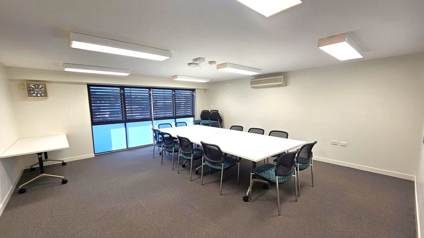 A conference room with a large rectangular table in the centre, surrounded by black chairs. On the left, there's a small table with wheels. The back wall features windows with horizontal blinds, and a stack of additional chairs is visible. The ceiling has several rectangular fluorescent lights, and an air conditioning unit is mounted on the right wall. The floor is carpeted in a grey tone.