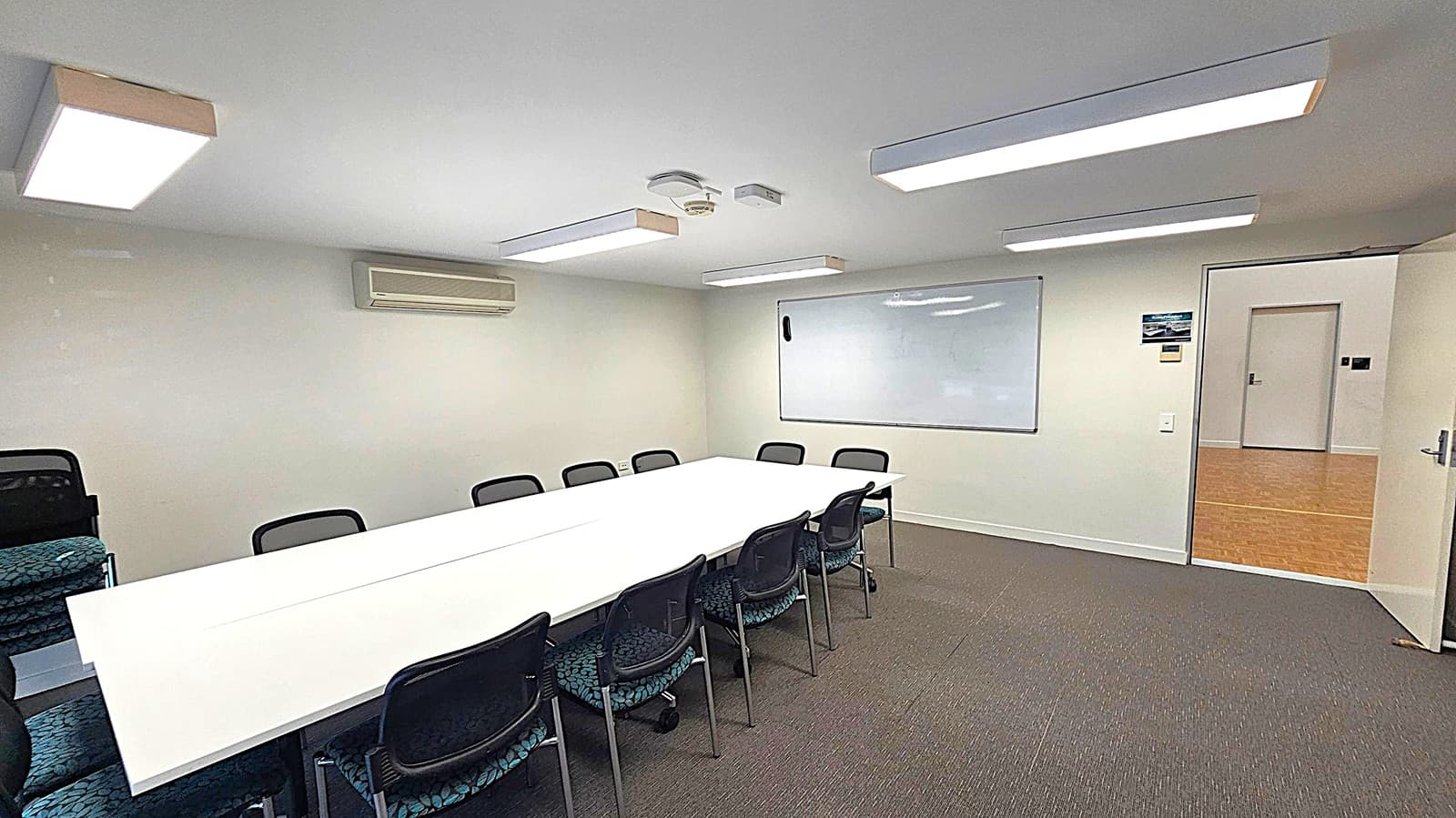 A conference room with a long white table in the centre surrounded by black chairs. On the left, there are additional stacked chairs. The back wall features a whiteboard and an air conditioning unit. The ceiling has rectangular fluorescent lights and a smoke detector. The floor is carpeted in grey. On the right, an open door leads to a hallway with a wooden floor.