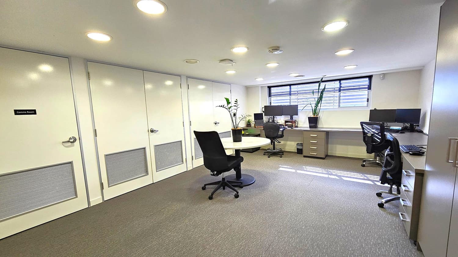 Office with grey carpet and white ceiling featuring recessed lights. On the left, two white doors, one labelled "Comms Room". In the centre, a round table with a black office chair and a potted plant. On the right, a long desk with multiple monitors, another office chair, and a potted plant. A window with horizontal blinds is above the desk. A cabinet is partially visible on the far right.