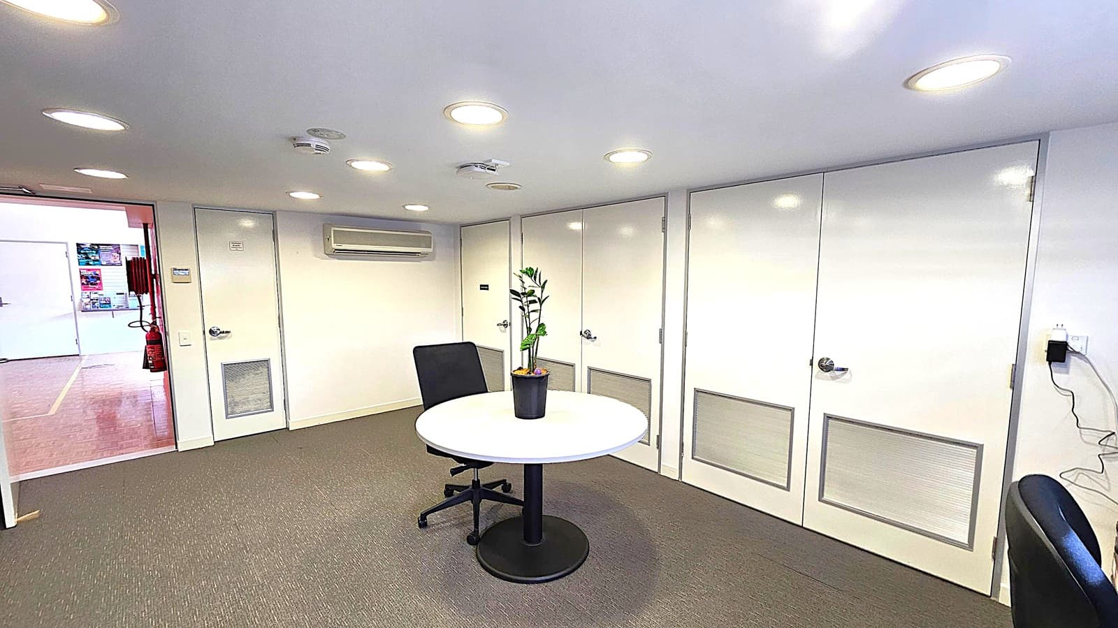 A small office space with a round table in the centre, featuring a potted plant. An office chair is positioned next to the table. The back wall has several white doors with ventilation panels and an air conditioning unit above. The floor is carpeted in a muted colour. The ceiling is fitted with recessed lights. On the left, an open doorway leads to another room with visible posters and a fire extinguisher. A power outlet with a cable is on the right wall.