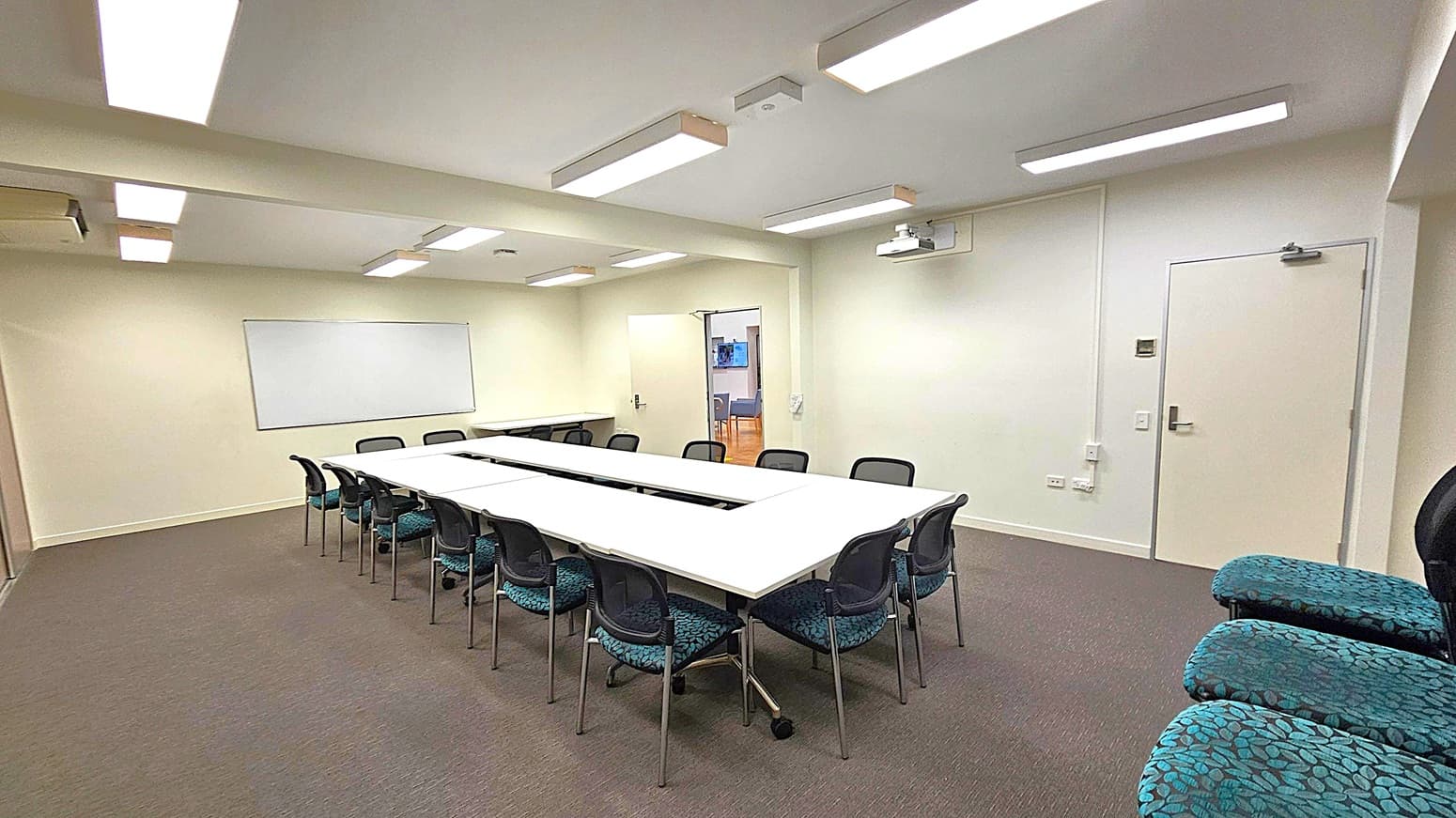 A conference room with a U-shaped arrangement of white tables surrounded by black chairs. On the left wall, a large whiteboard is mounted. The right wall has a closed door and a projector mounted on the ceiling. The floor is carpeted in grey, and the ceiling features several rectangular fluorescent lights. In the background, an open door leads to another room with visible chairs and a screen.