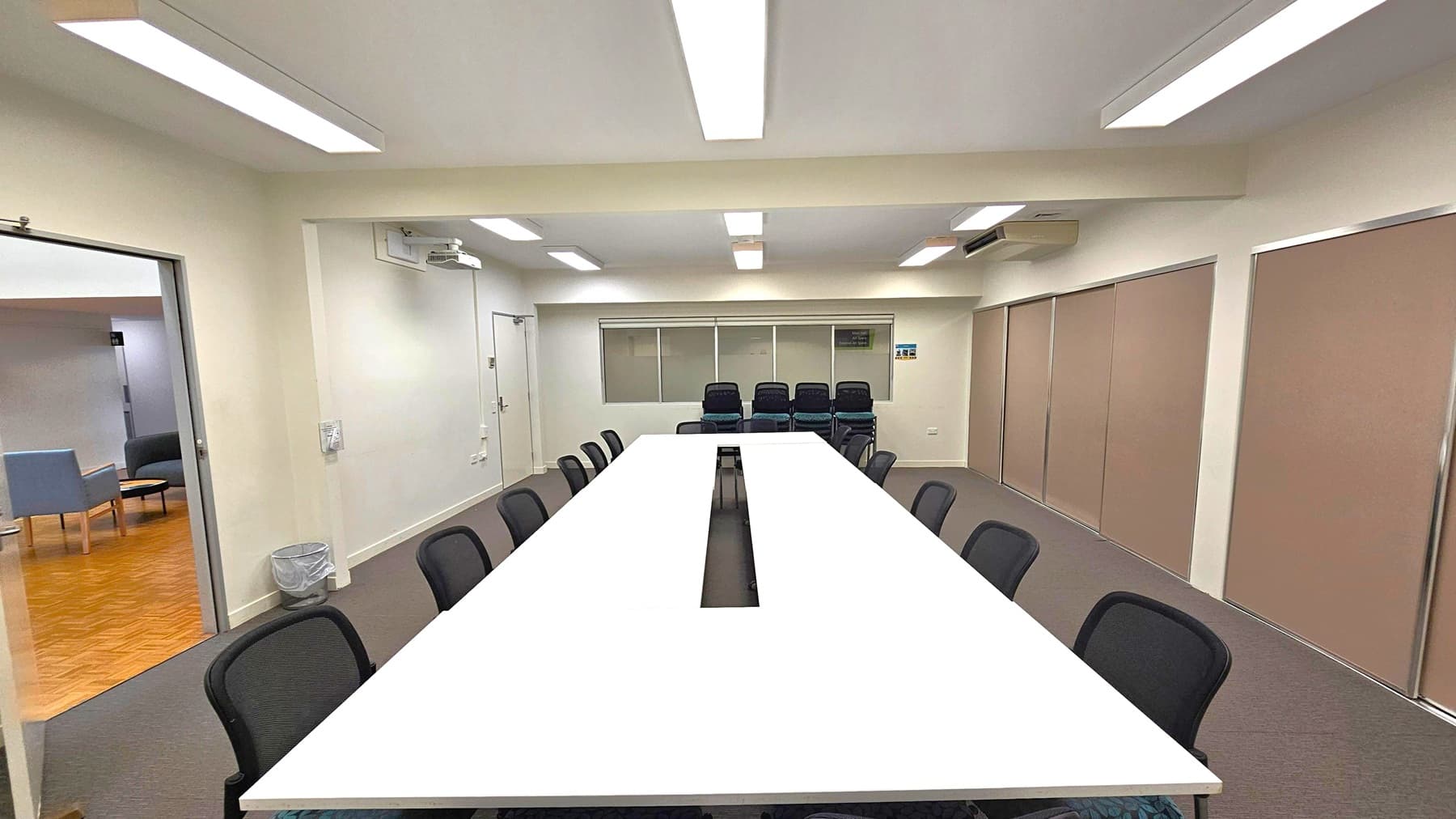 A conference room with a long, white rectangular table in the centre, surrounded by black chairs. The floor is carpeted in grey. On the left, an open door leads to another room with wooden flooring and a few chairs. The back wall has a row of windows and a stack of additional chairs. The right wall features large, beige sliding panels. The ceiling is fitted with fluorescent lights, and there is an air conditioning unit on the right wall.