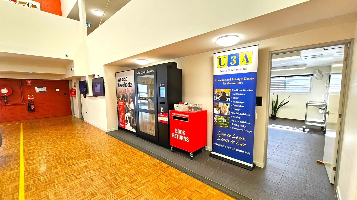 A hallway with a parquet floor and a white ceiling. On the left, a red wall with a fire hose reel and extinguisher. In the centre, a book vending machine and a red book return bin. To the right, a blue and yellow U3A banner advertising classes for those over 50. An open door leads to a room with grey tiles and a plant near the window.