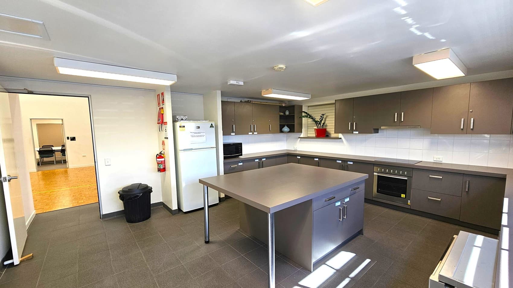 A modern kitchen with grey tiled flooring and a white ceiling featuring rectangular lights. In the centre, a large stainless steel island with drawers. The right side has dark cabinets, a built-in oven, and a white tiled backsplash. A plant in a red pot sits on the counter. On the left, a white fridge and a microwave. A fire extinguisher and a black bin are near the open door, which leads to a room with wooden flooring and chairs.