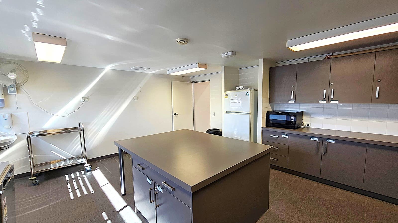 A kitchen with a central island featuring drawers and a smooth countertop. On the right, there are dark cabinets with a microwave on the counter. A white refrigerator is next to the cabinets. On the left, a metal trolley with wheels is near the wall, which has a mounted fan. The floor is tiled in a dark colour, and the ceiling has rectangular fluorescent lights. Sunlight creates patterns on the floor and walls.