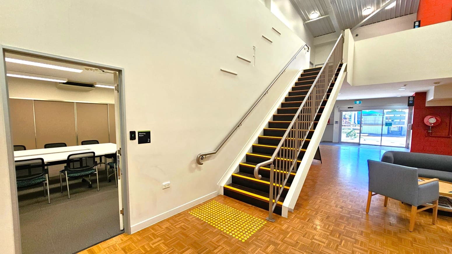 On the left, there's an open door leading to a meeting room with a long table and several chairs. The floor is carpeted inside the room. In the centre, a staircase with black steps and yellow edges ascends to the upper level, with a metal handrail on the right. The floor is wooden parquet. On the right, there's a seating area with grey chairs and a table. In the background, large glass doors provide a view of the outside, and a red wall features a fire hose reel. The ceiling is high with recessed lighting.