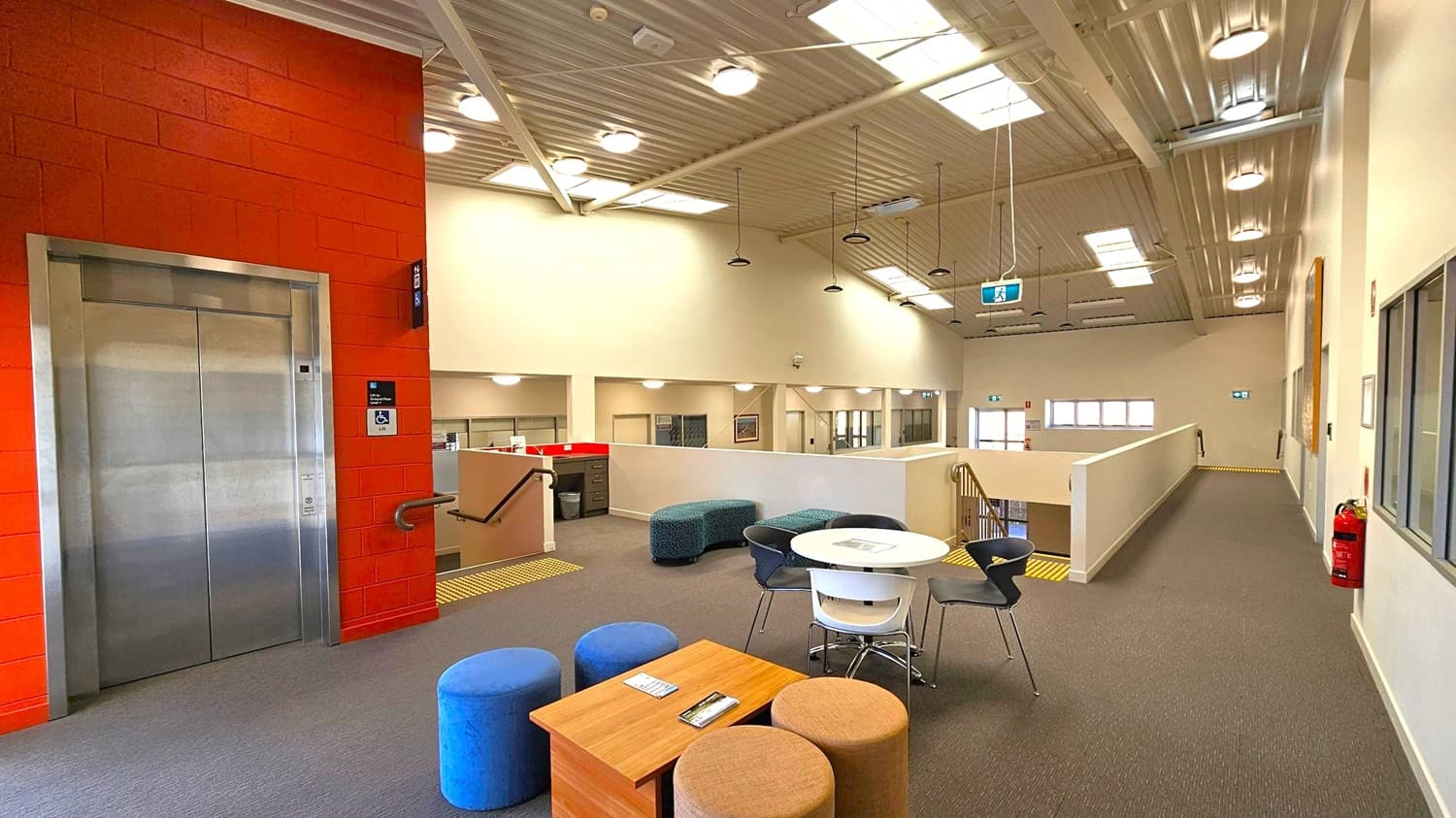 A modern indoor space with a grey carpeted floor. On the left, there's a silver lift set in a red brick wall. In the centre, a seating area features a round white table surrounded by black and white chairs, and several colourful pouffes. A wooden table holds some pamphlets. The right side has a corridor with a fire extinguisher on the wall. The ceiling is high with exposed beams and multiple lights, and a staircase is visible in the background.