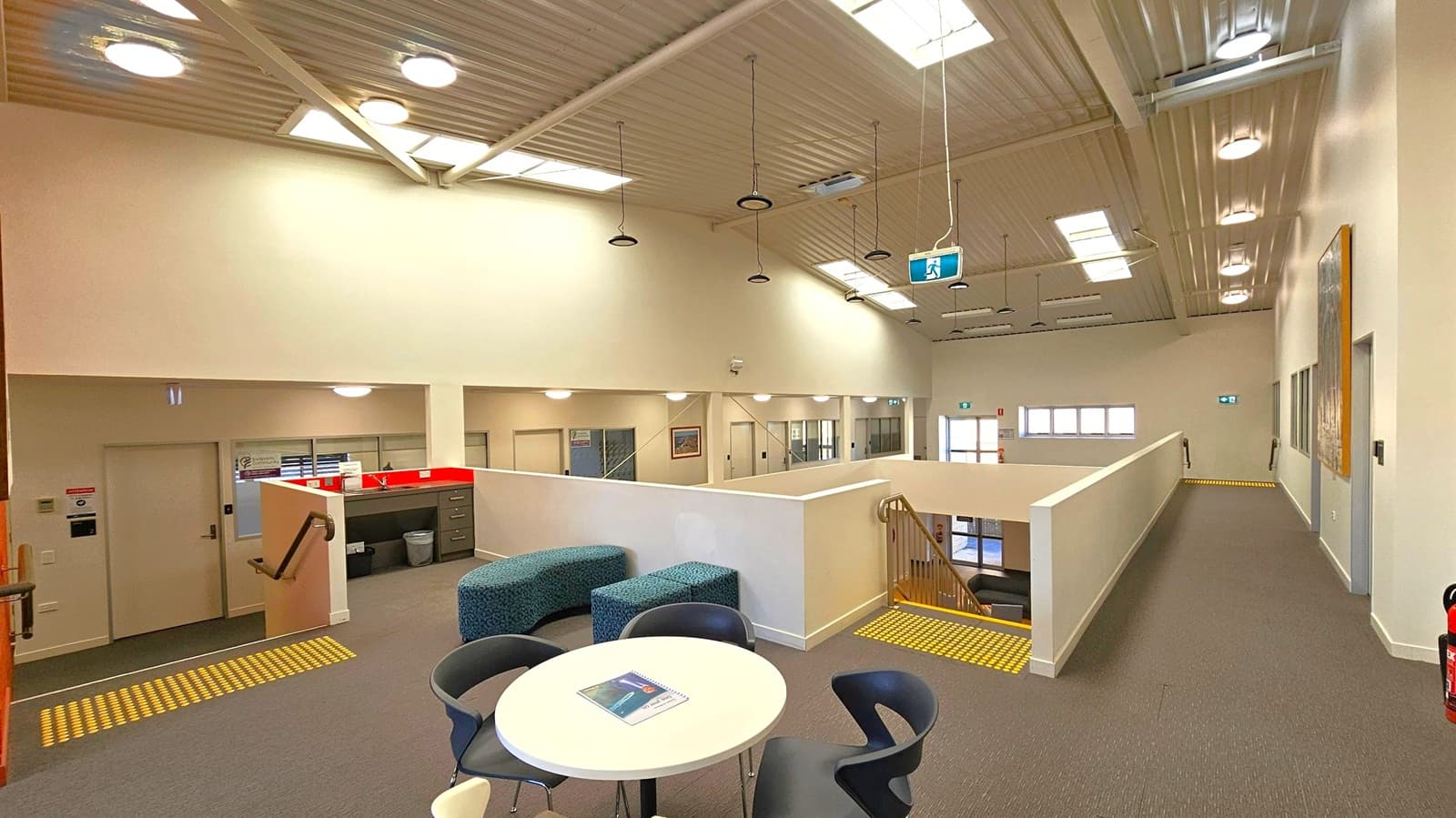 A spacious interior with a high, corrugated metal ceiling featuring multiple skylights and hanging lights. In the foreground, a round table with chairs and a notebook is visible. To the left, a small kitchenette area with a sink and cabinets is adjacent to a staircase with a handrail. The right side has a long corridor with doors and a fire extinguisher. The floor is carpeted, and there are yellow tactile indicators near the stairs. Walls are light-coloured, enhancing the bright atmosphere.