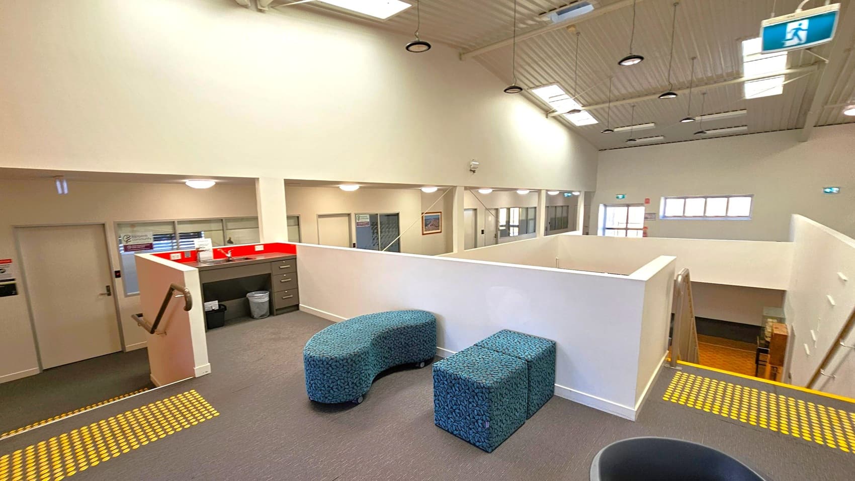 A spacious interior with a high ceiling featuring skylights and hanging lights. In the foreground, there are two blue, patterned seating units on a grey carpet. To the left, a small kitchenette with a red splashback and cabinets. A staircase with a handrail descends to the lower level. The background shows a corridor with multiple doors and windows, and a few framed pictures on the walls. An emergency exit sign is visible near the ceiling.