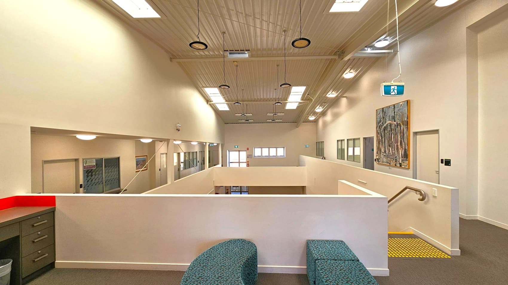 A spacious interior with a high, wooden-panelled ceiling featuring multiple skylights and hanging lights. The foreground has a curved, patterned bench and a green cabinet with drawers. The background shows a corridor with doors and windows, a large painting on the right wall, and an exit sign hanging from the ceiling. The floor is carpeted, and a staircase with a yellow safety strip is visible on the right.